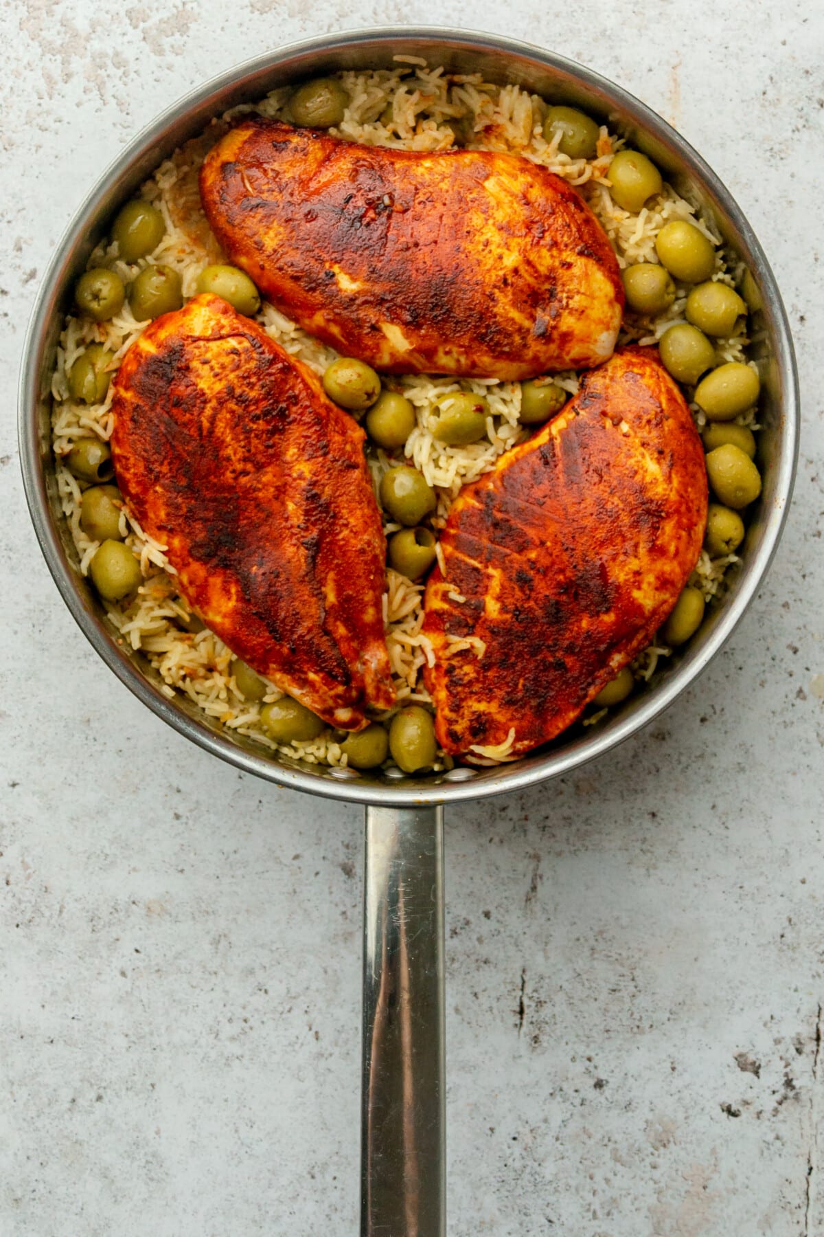 Spiced cooked chicken breasts and green olives sit on top of rice in a stainless steel frying pan on a light grey surface.