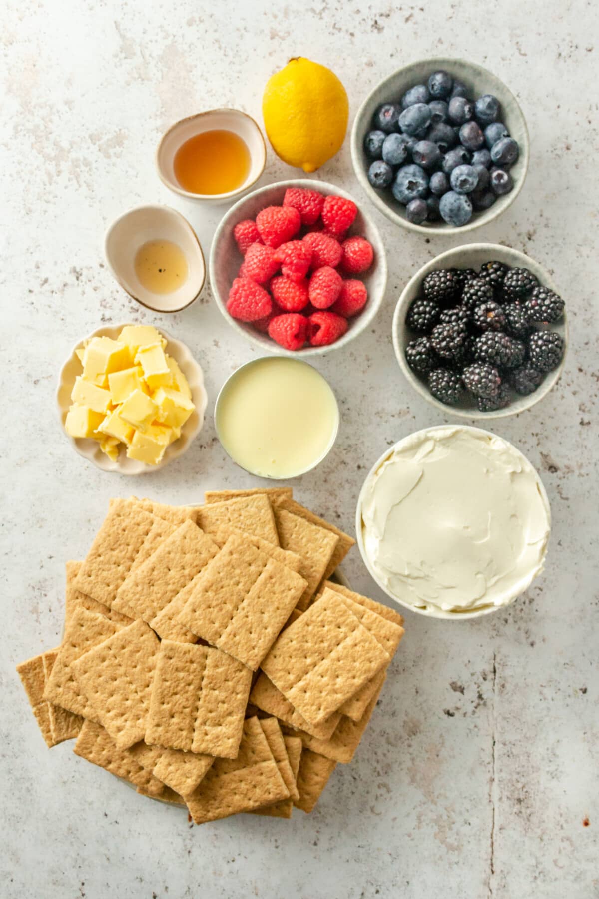 Ingredients for no bake cheesecake bars sit in a variety of bowls on a light grey surface.