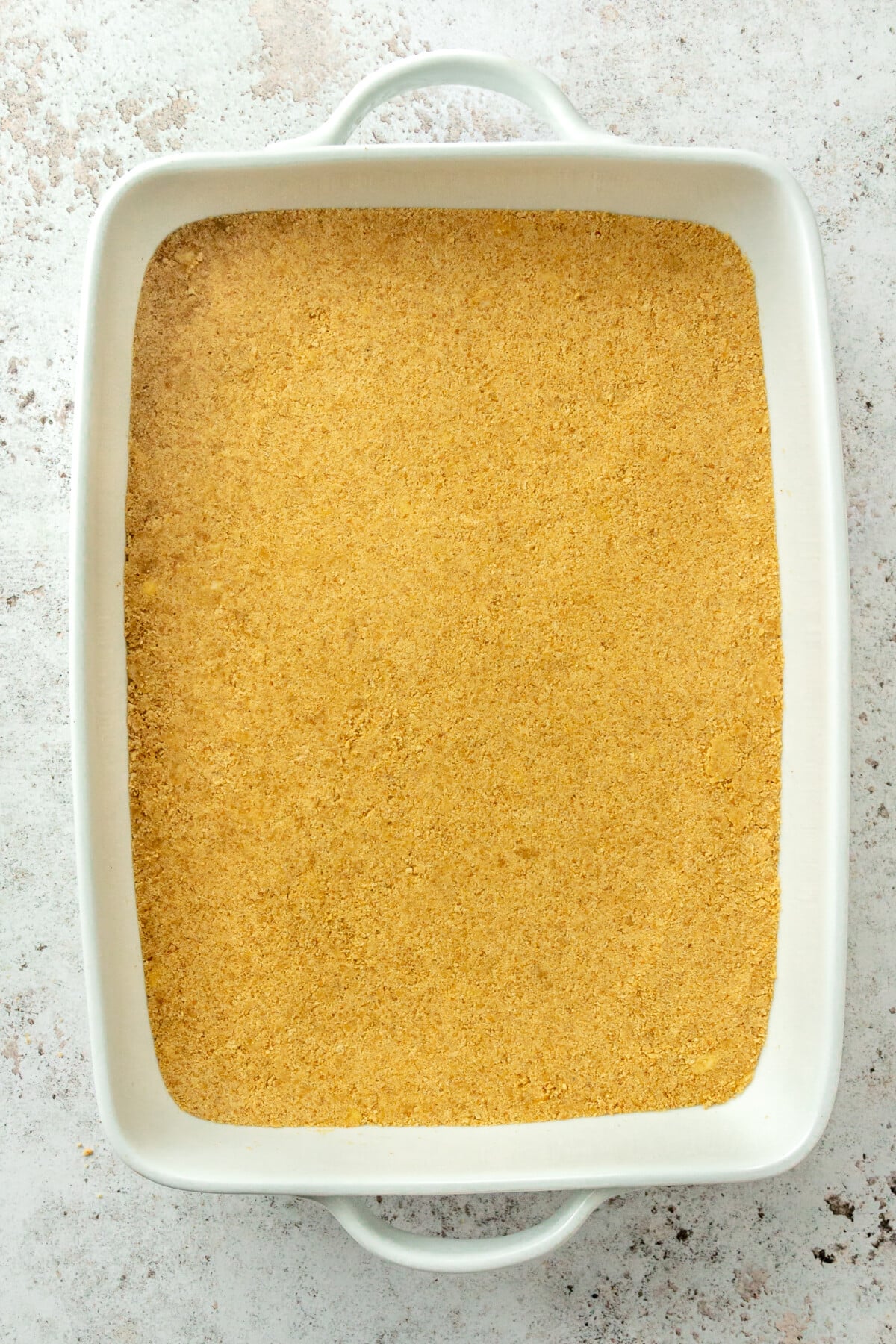 The graham cracker mixture is shown pressed into an even layer in the bottom of a white rectangular baking dish.