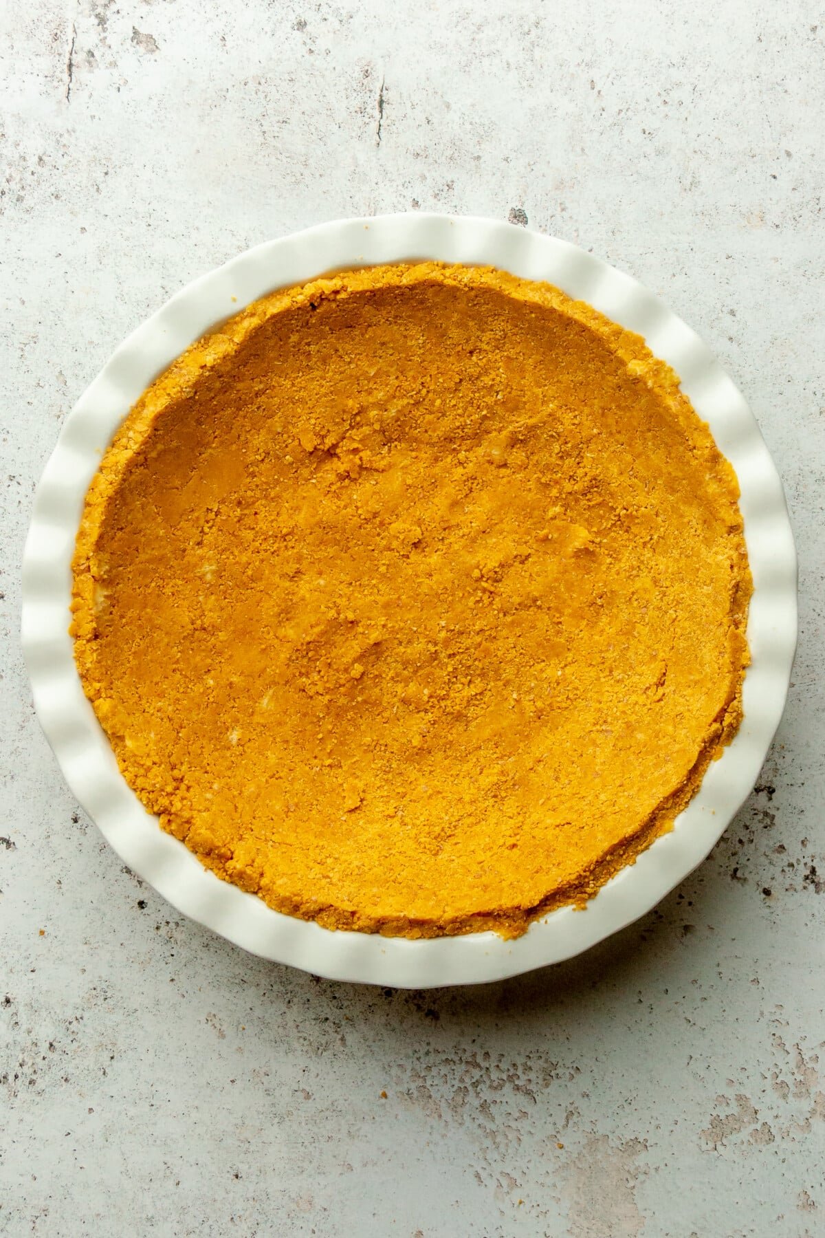 A crushed graham cracker crust sits in a white ceramic pie dish on a light gray surface.