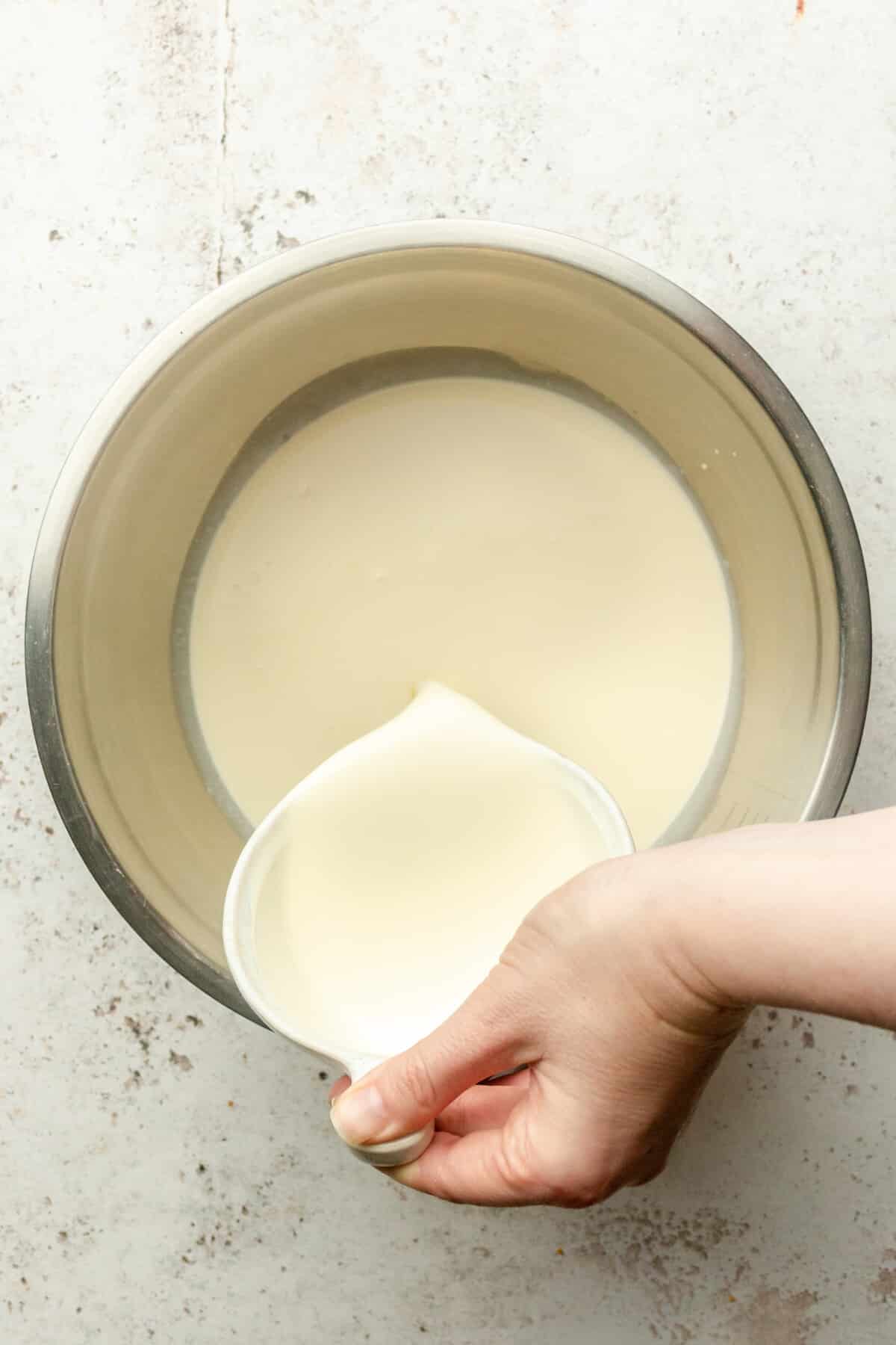 Heavy cream is poured into a stainless steel bowl on a light gray surface.