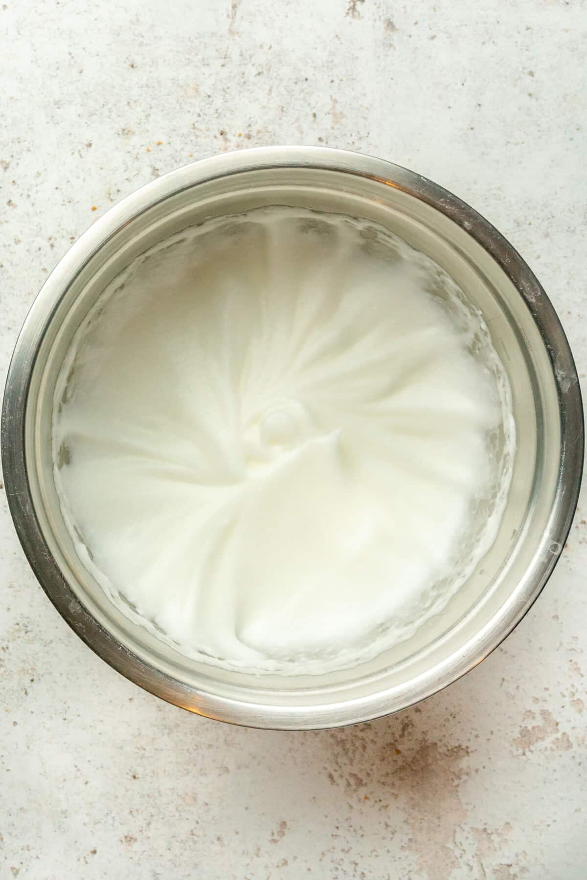 Stiff whisked egg whites sit in a stainless steel bowl on a light gray surface.