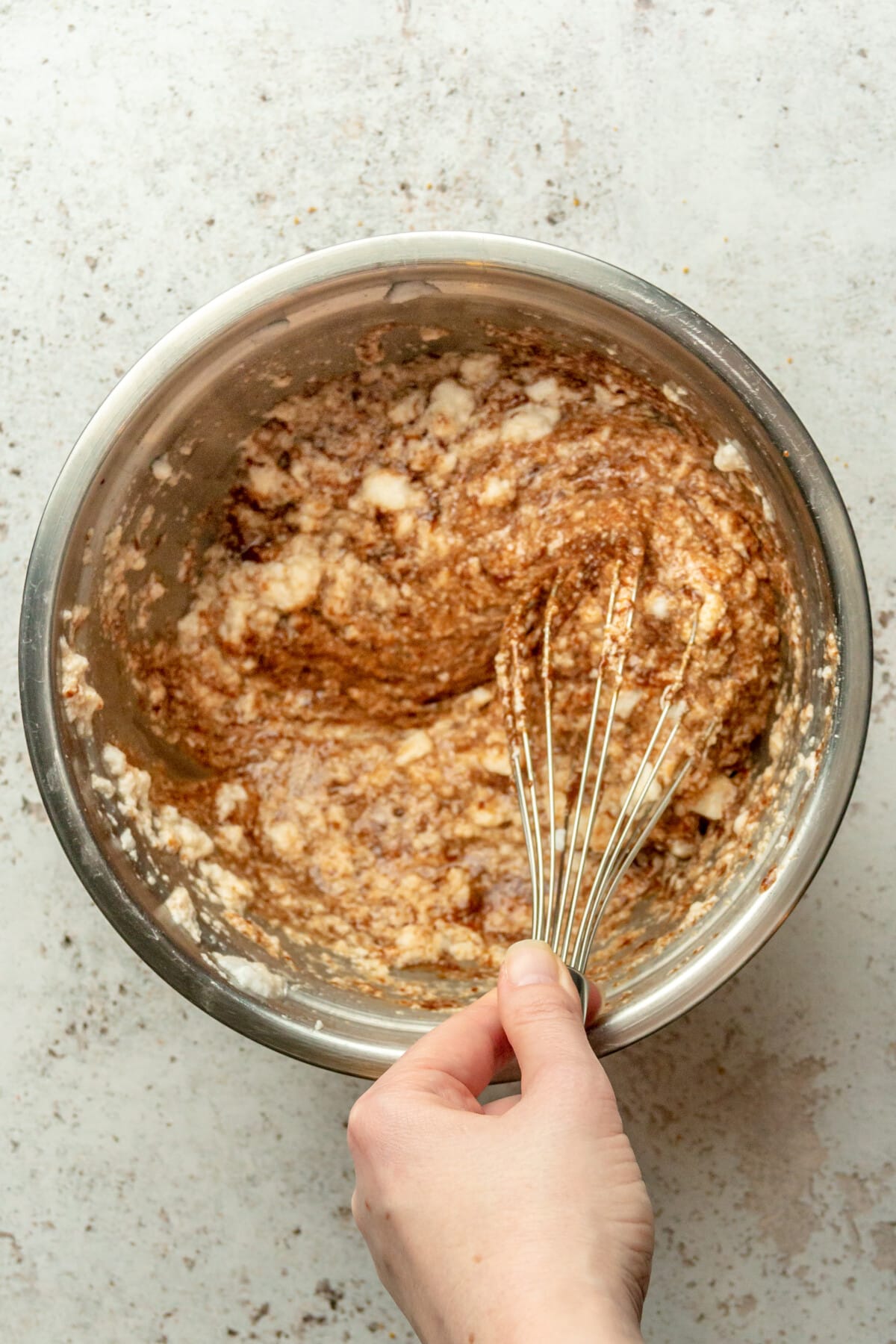 A melted chocolate and egg yolk mixture is folded through whipped egg whites in a stainless steel bowl on a light gray surface.