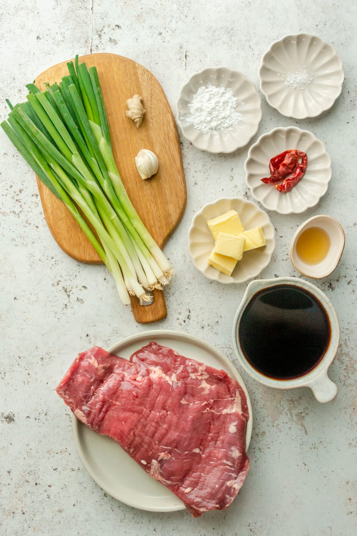 Ingredients for paleo Mongolian beef sit in a variety of bowls and plates on a light grey surface.