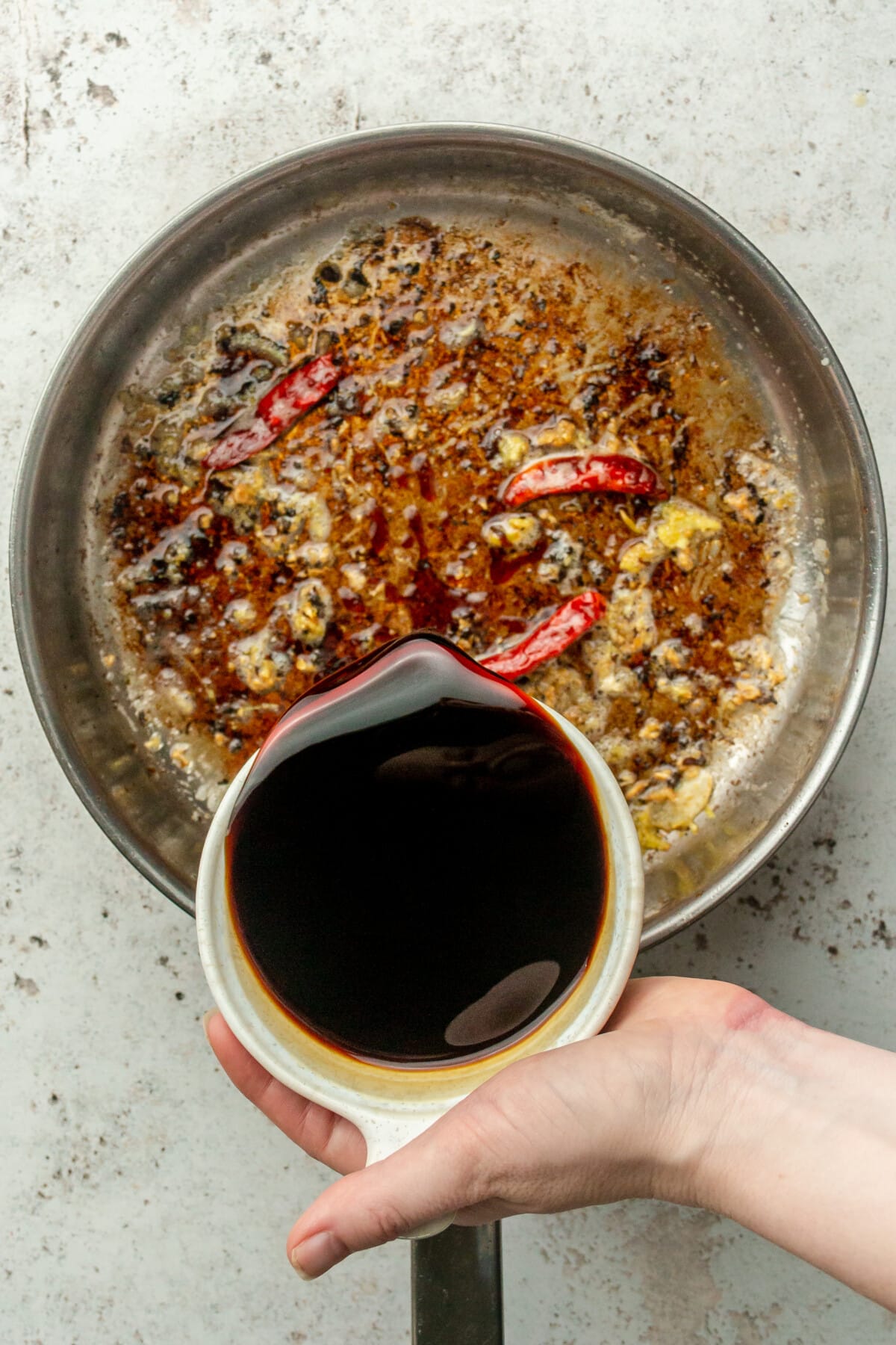 Coconut aminos are poured over garlic, ginger and dried chilis in a stainless steel saucepan on a light gray surface.