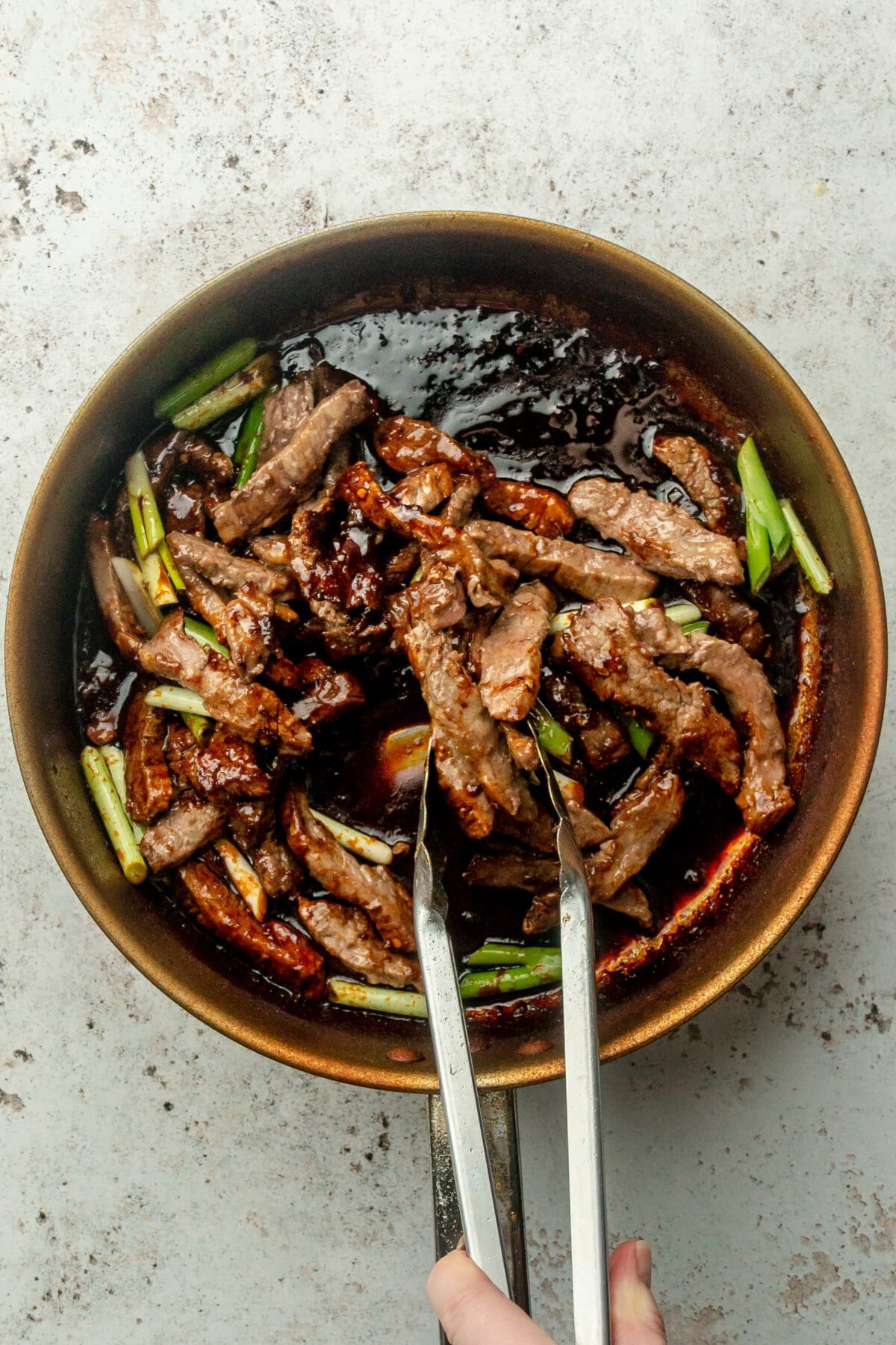 Pieces of beef are tosed through a Mongolian sauce in a stainless steel saucepan on a light gray surface.