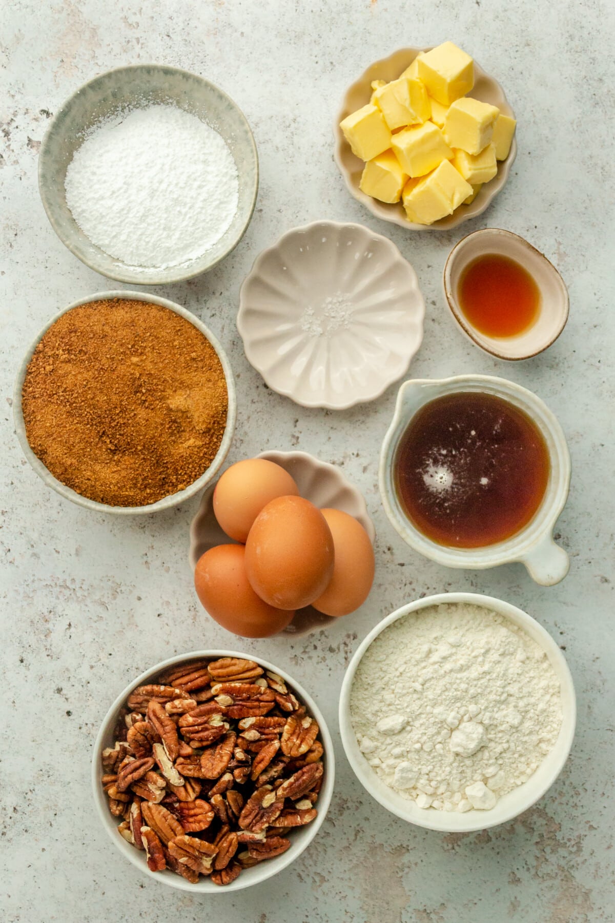 Ingredients for pecan pie bars sit in a variety of bowls and plates on a light grey surface.