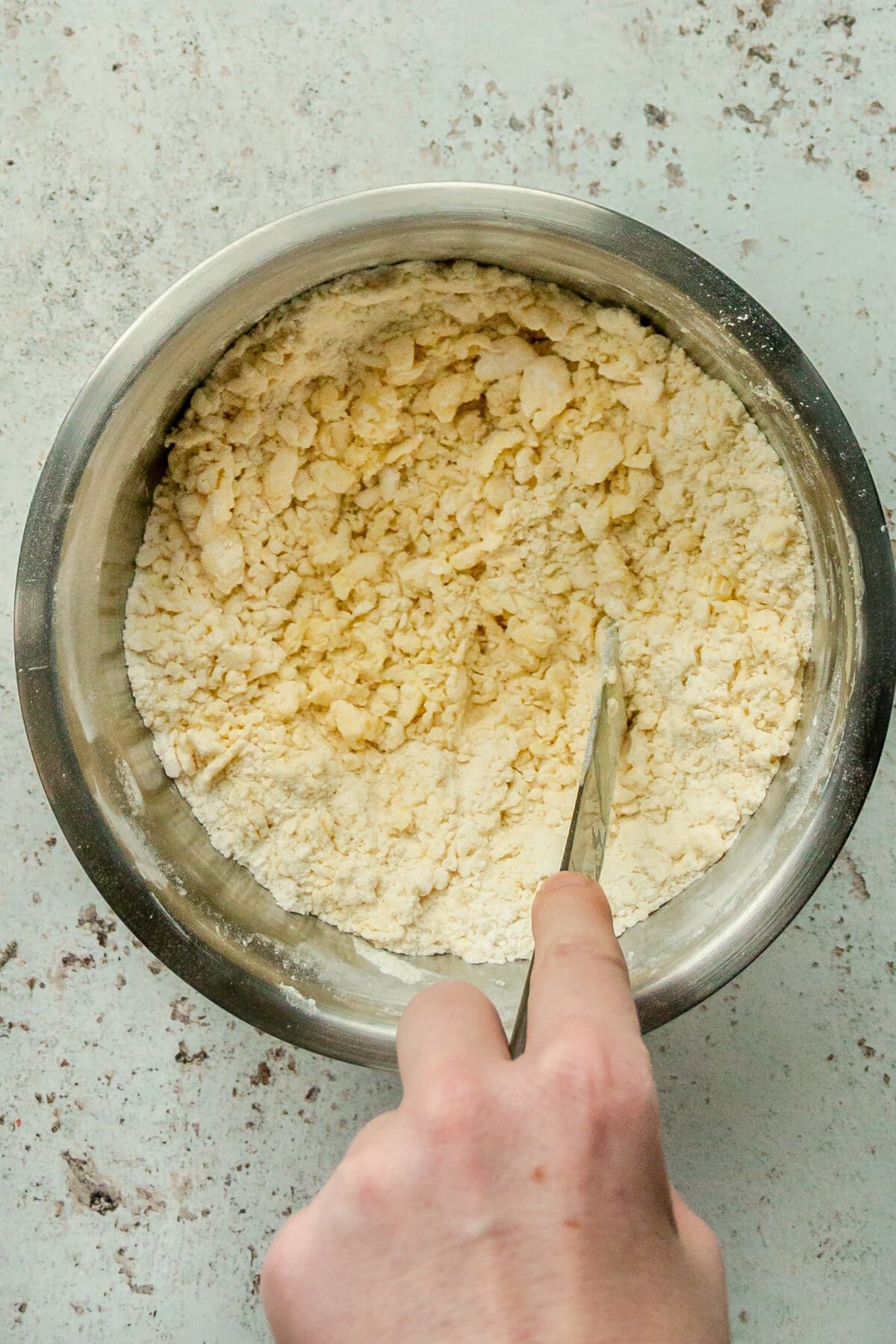 Butter is cut into a flour and sugar mixture in a stainless steel bowl on a light gray surface.