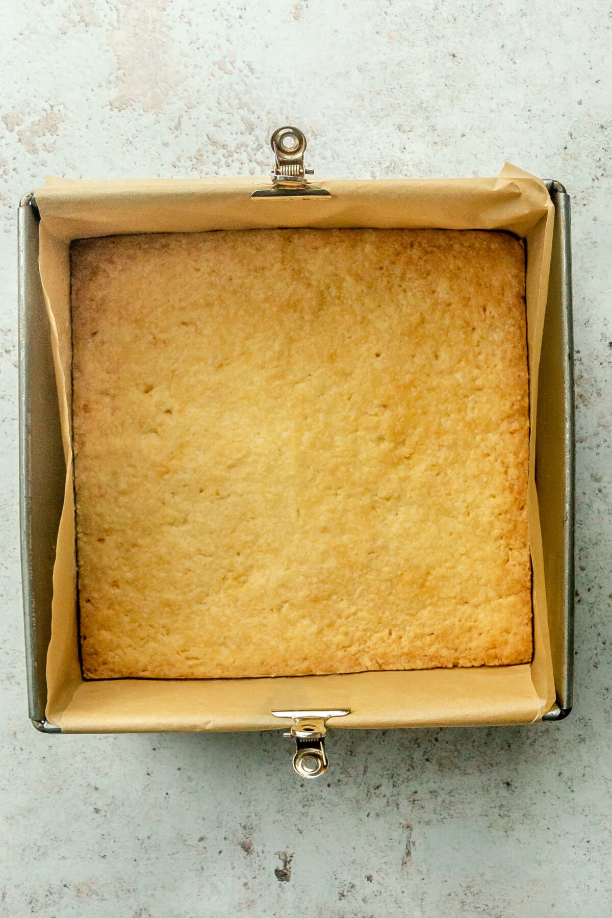 Baked shortbread crust sits in a parchment lined square metal tin on a light gray surface.