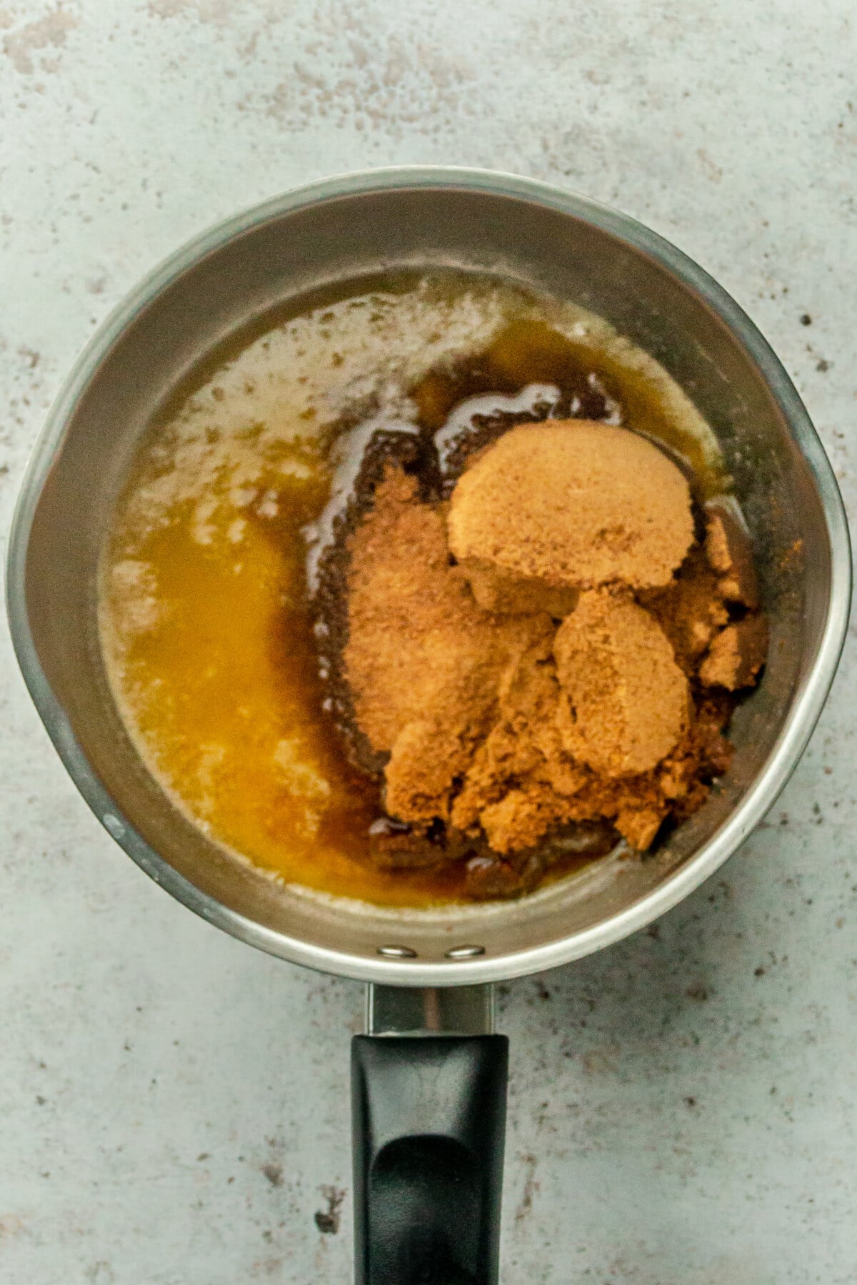 Coconut sugar sits in a melted butter in a stainless steel saucepan on a light gray surface.