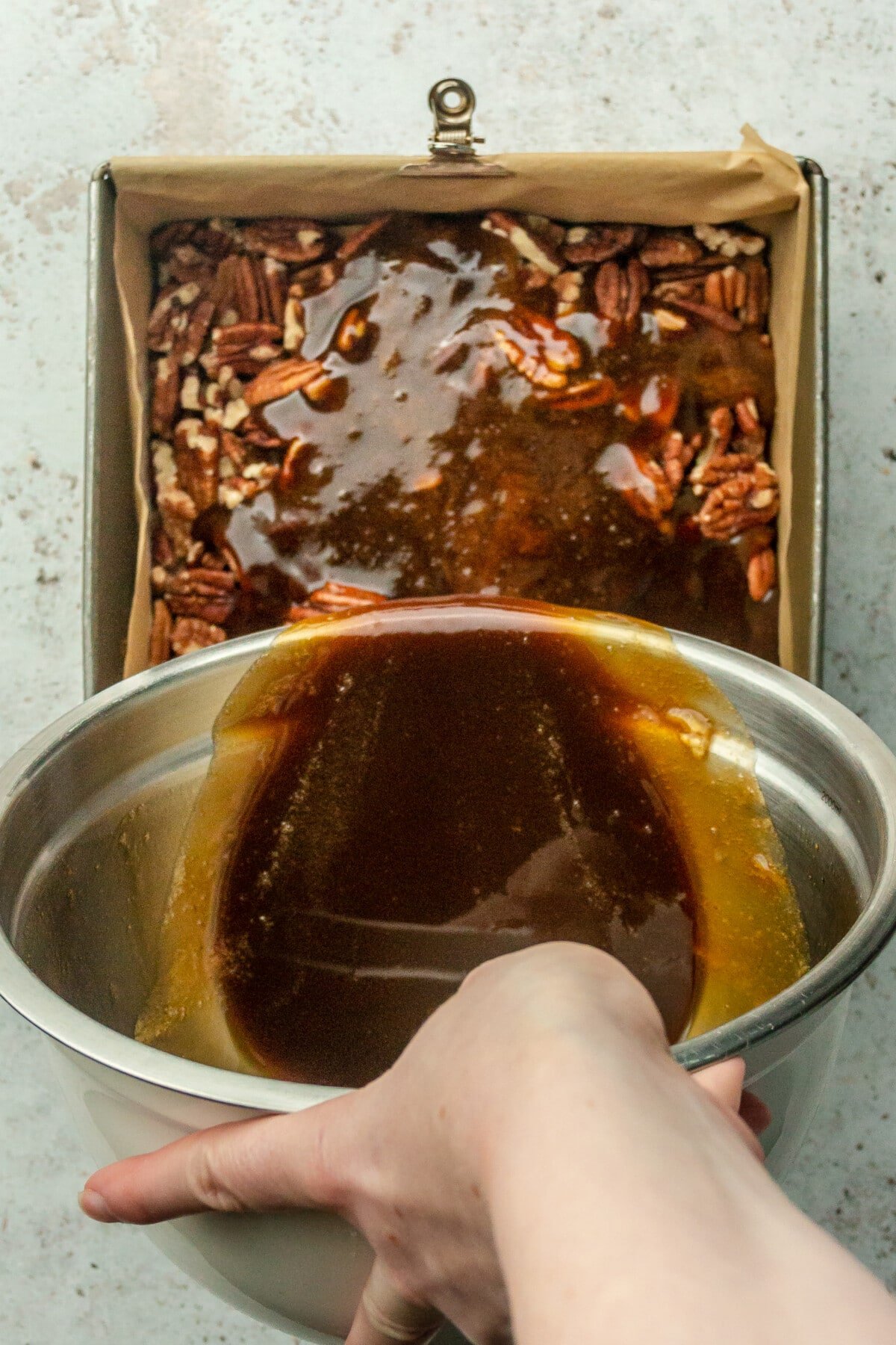 A pecan pie mixture is poured over pecans in a parchment paper lined baking tin on a light gray surface.