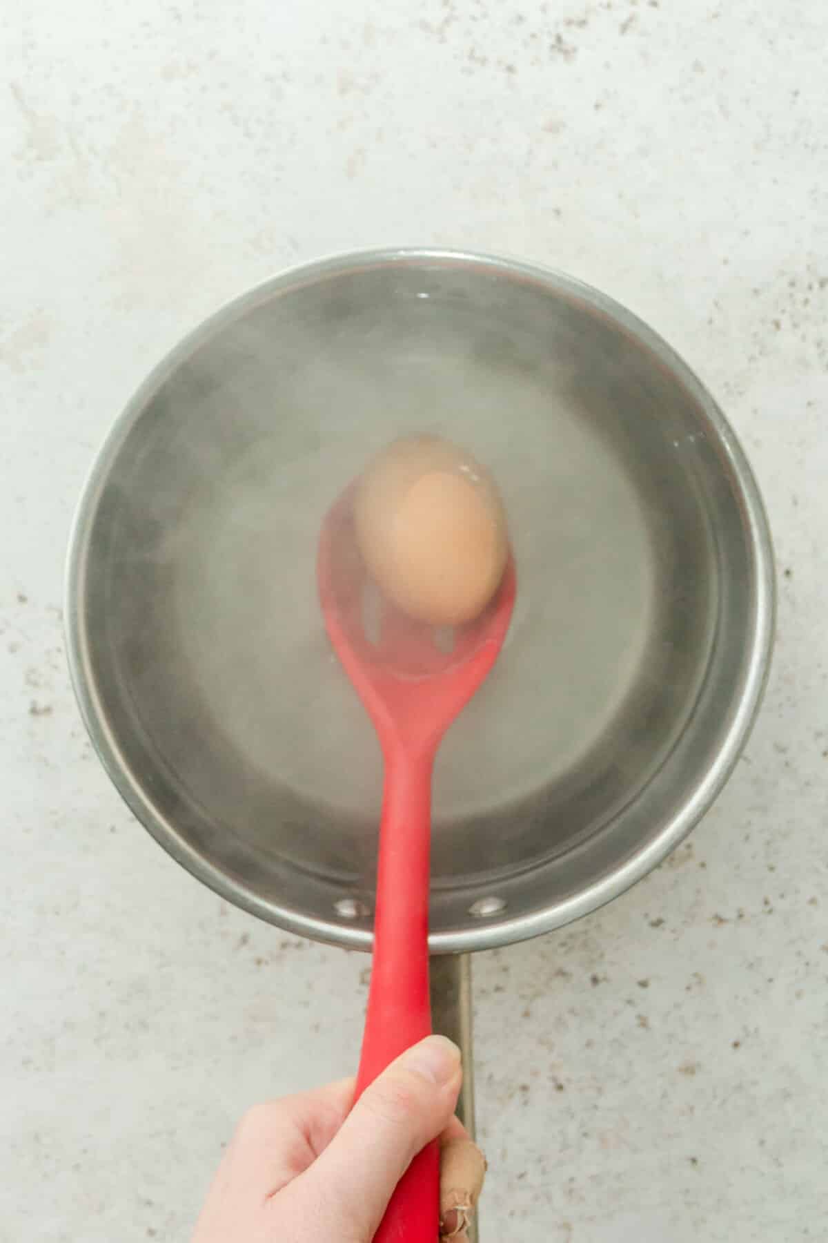 An egg is dropped into hot water in a stainless steel saucepan on a gray surface.