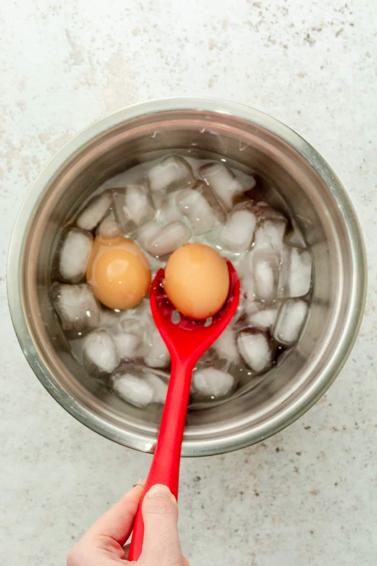 An egg is placed into a stainless steel bowl of ice water on a gray surface after being boiled.