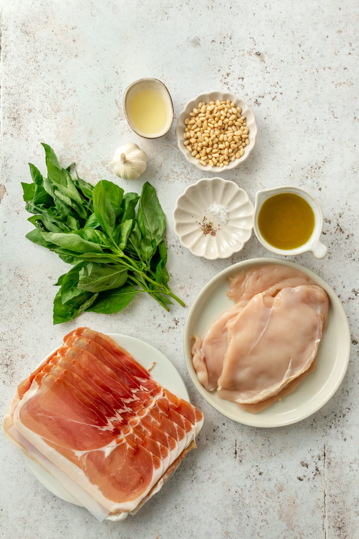 Ingredients for prosciutto chicken breasts sit in a variety of bowls on a light grey surface.