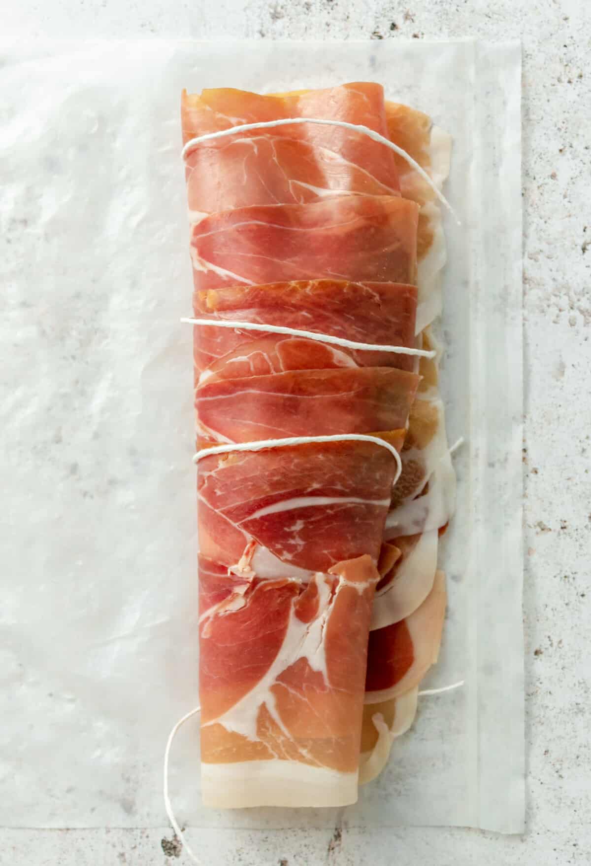 A piece of cooking string is shown aiding in wrapping the prosciutto around the chicken breast.