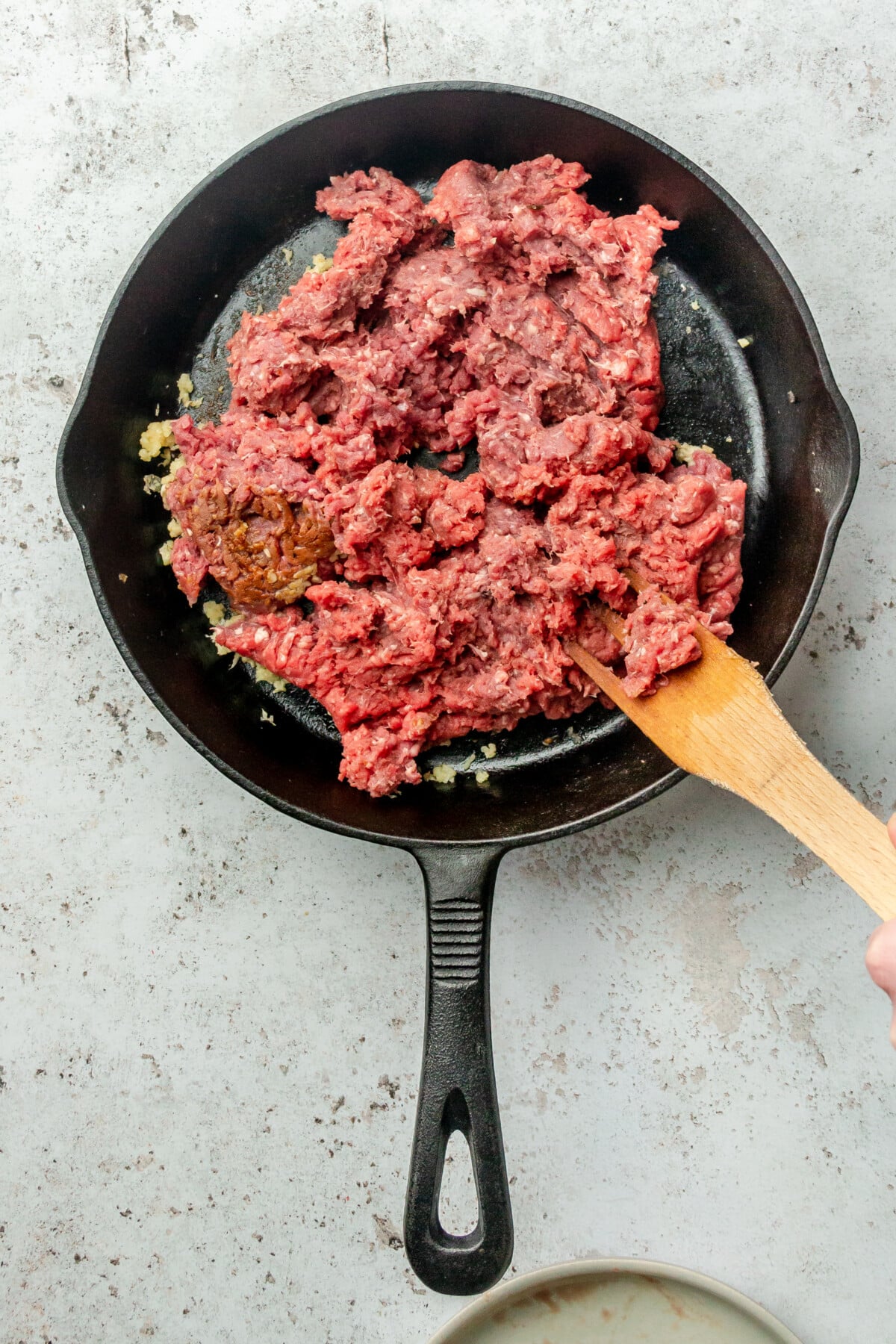 Minced beef is stirred in a cast iron skillet on a light grey colored surface.