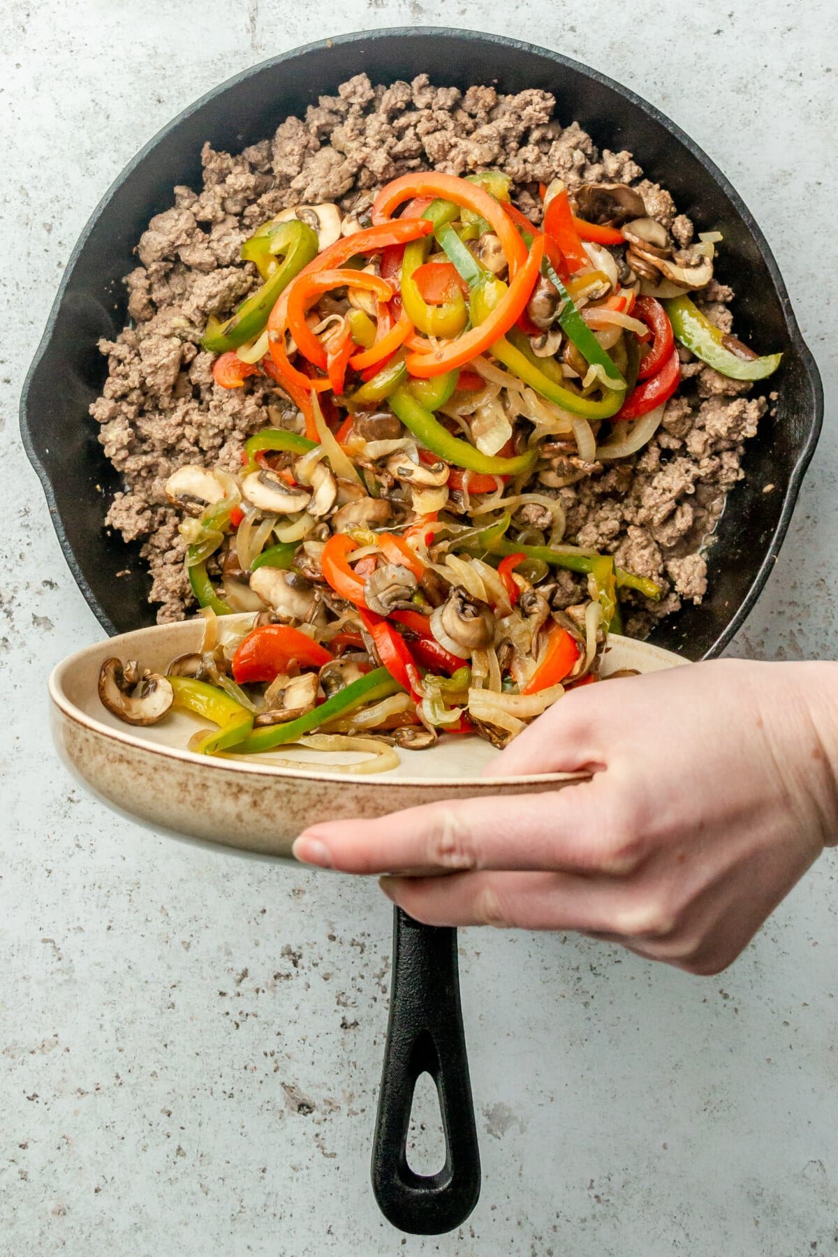Cooked vegetables are tossed into browned minced beef in a cast iron skillet sitting on a light grey colored surface.