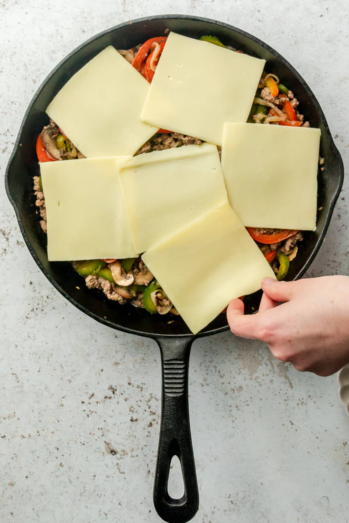 Slices of cheese are laid over minced beef and vegetables in a cast iron skillet on a light grey colored surface.