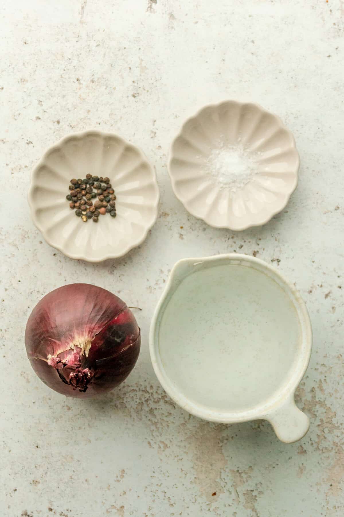 Ingredients for pickled red onions sit in a variety of bowls and plates on a light grey surface.