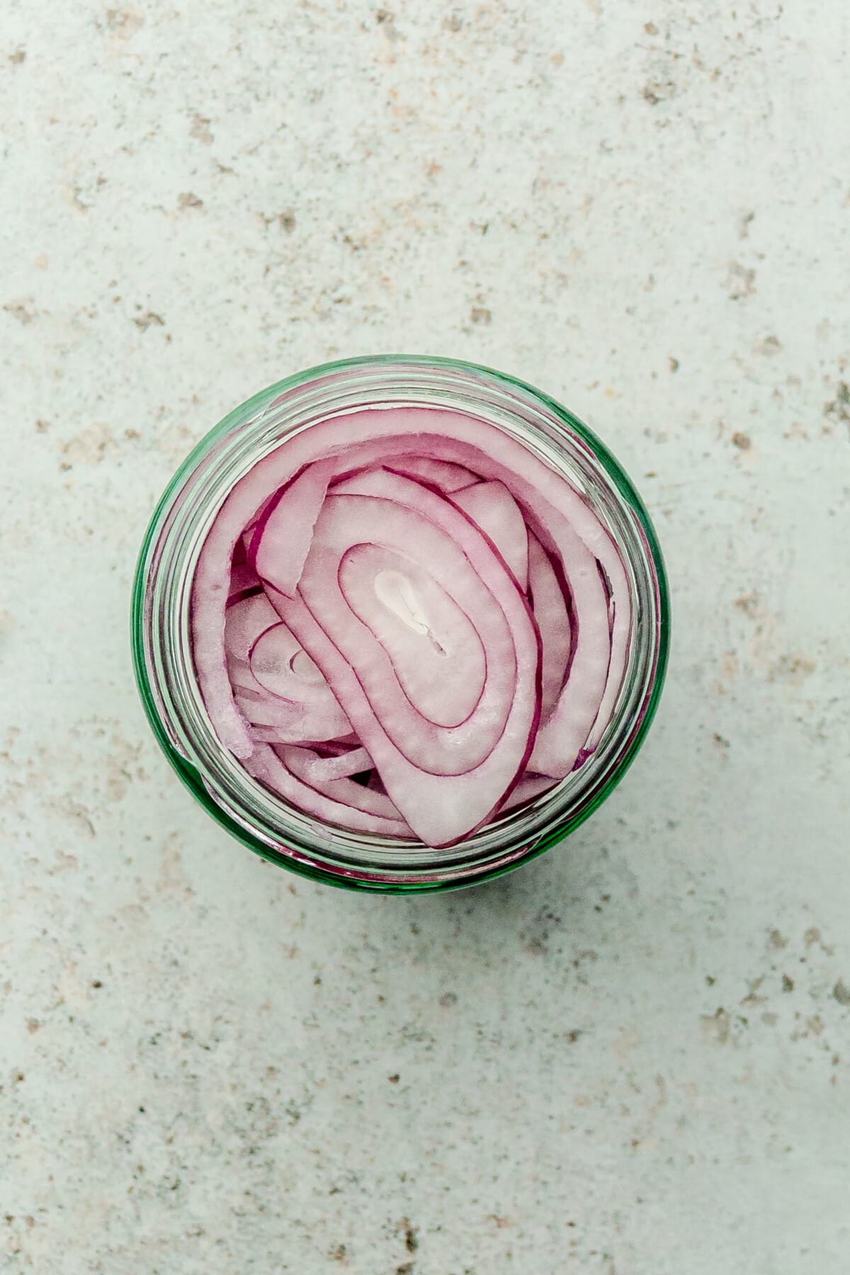 Sliced red onions are sitting in a glass jar on a light gray surface.
