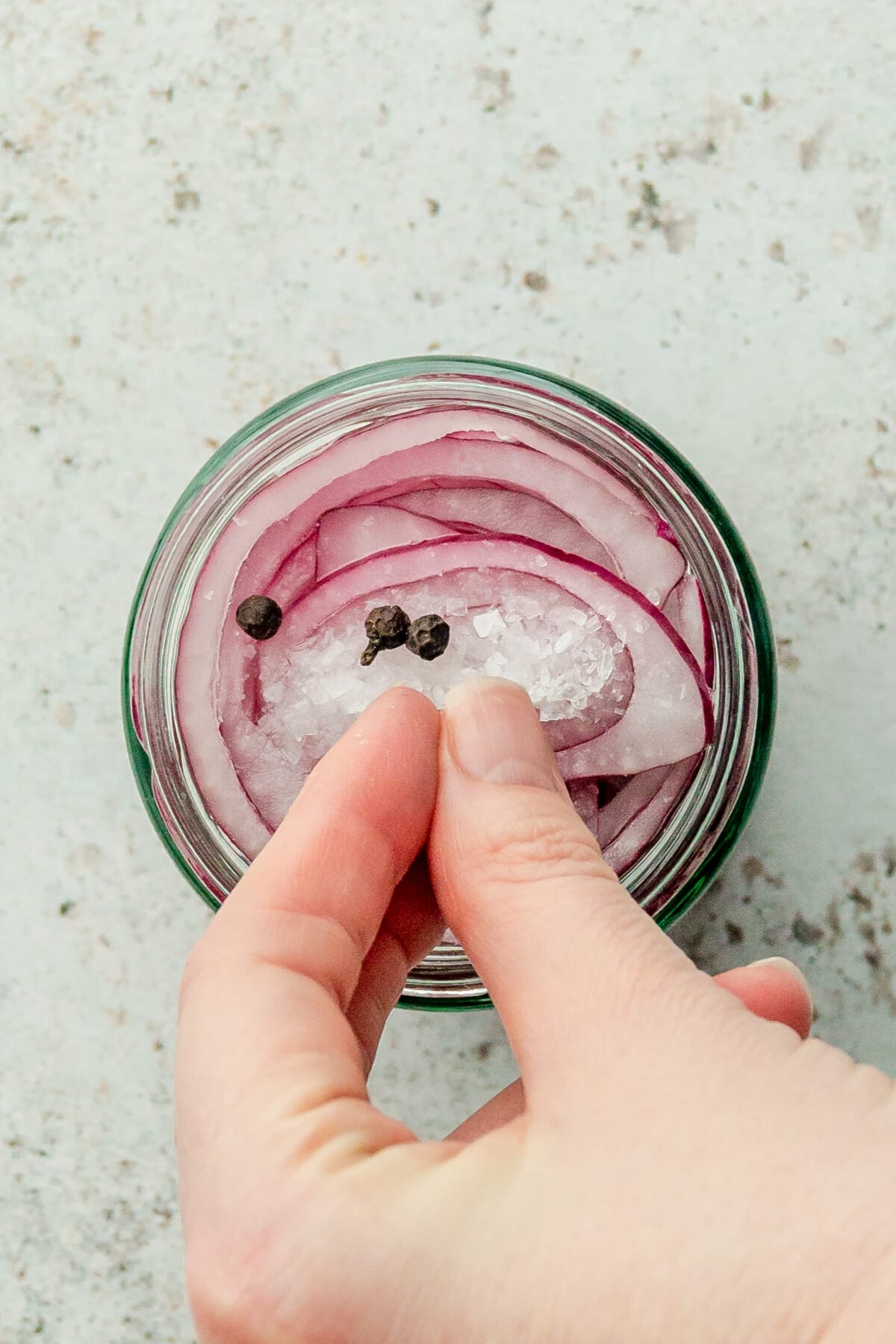Whole peppergrains are sprinkled over sliced red onions and salt in a glass jar with vinegar on a light gray surface.
