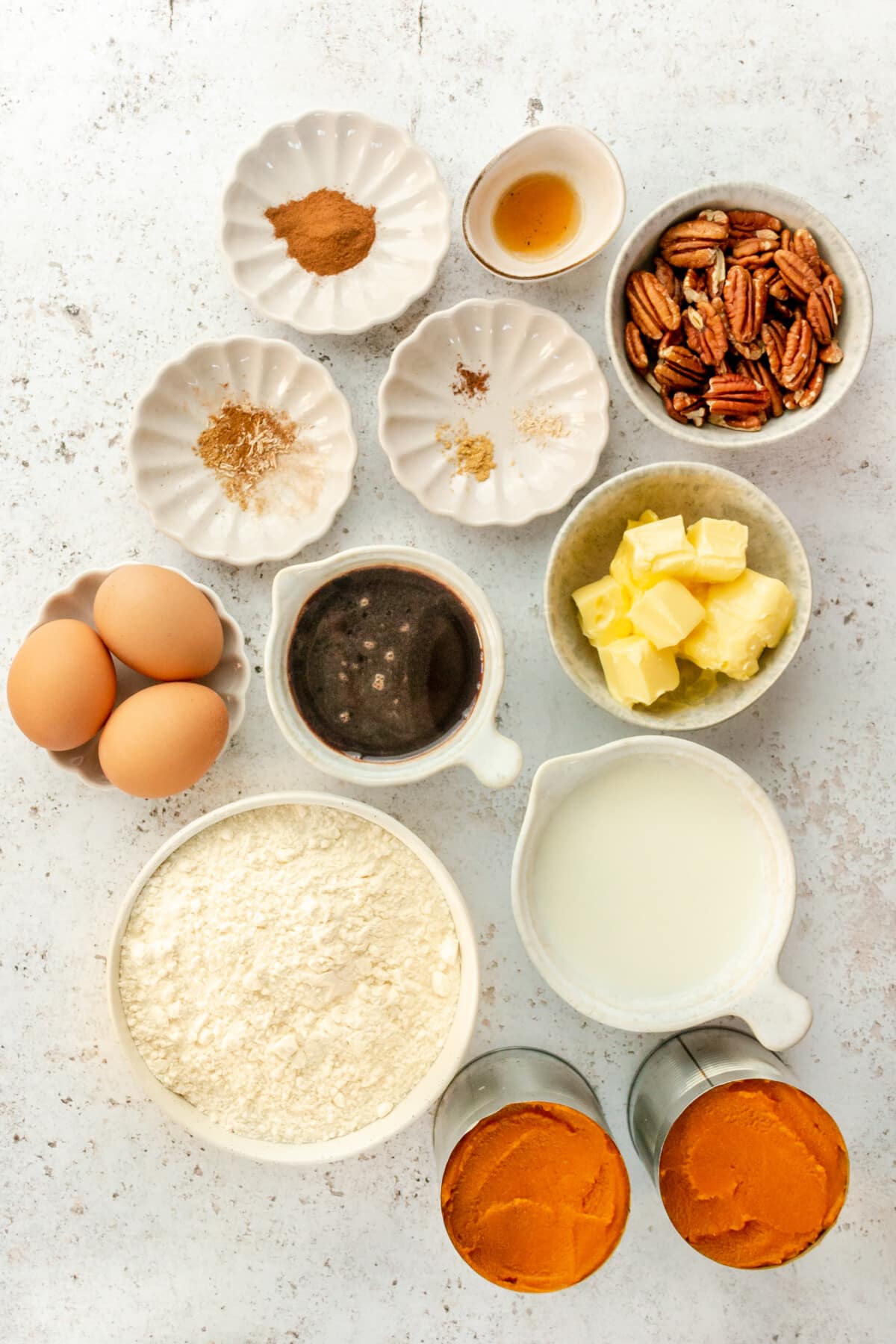 Ingredients for pumpkin dump cake sit in a variety of bowls and plates on a light grey surface.