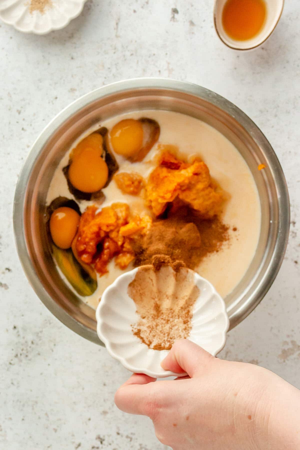 Spices are tossed into a pumpkin cake batter sitting in a stainless steel bowl on a light grey surface.