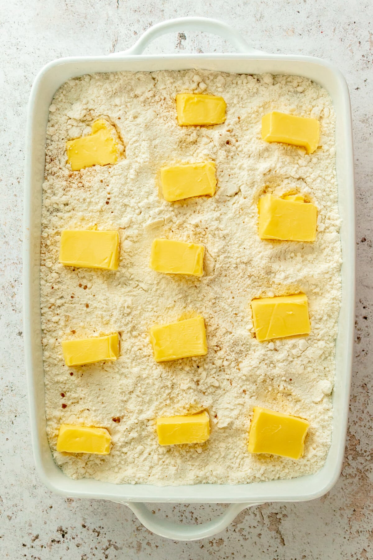 Slices of butter sit on top of a pumpkin dump cake mixture in a white rectangle baking dish on a light grey surface.