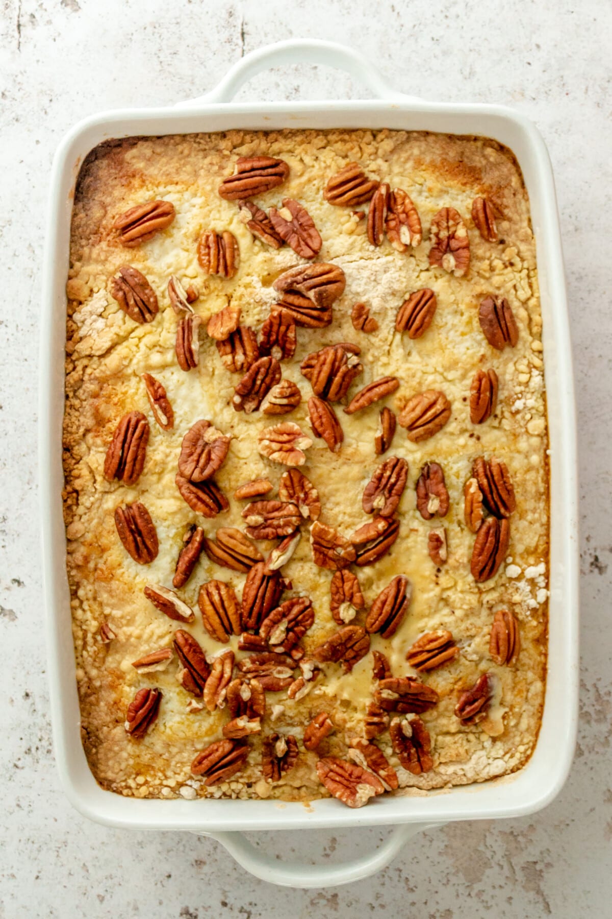 Pecans sit on top of a pumpkin dump cake on a white rectangle baking dish on a light grey surface.