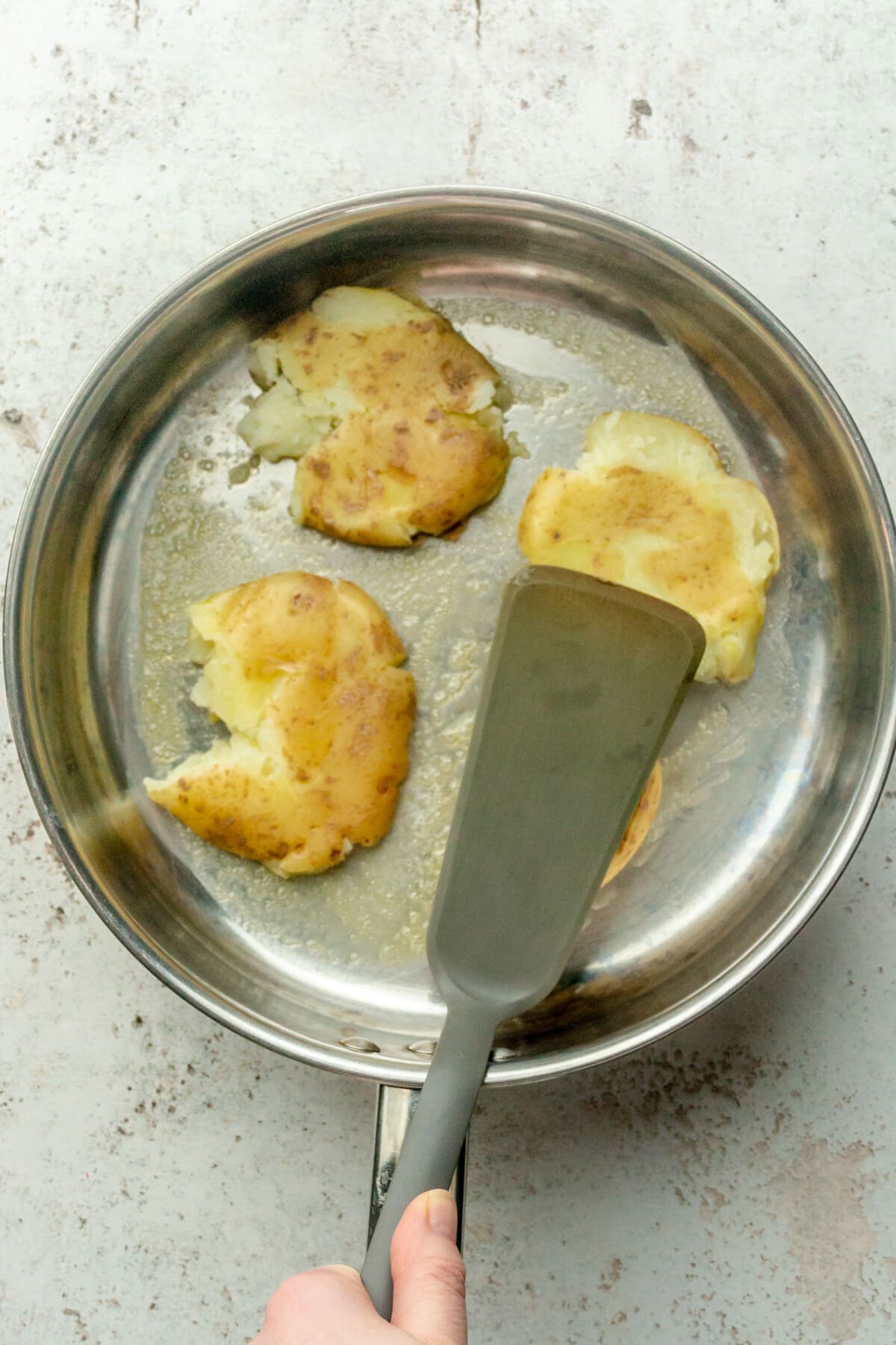 A spatula smashes potatoes in a stainless steel saucepan on a light gray surface.