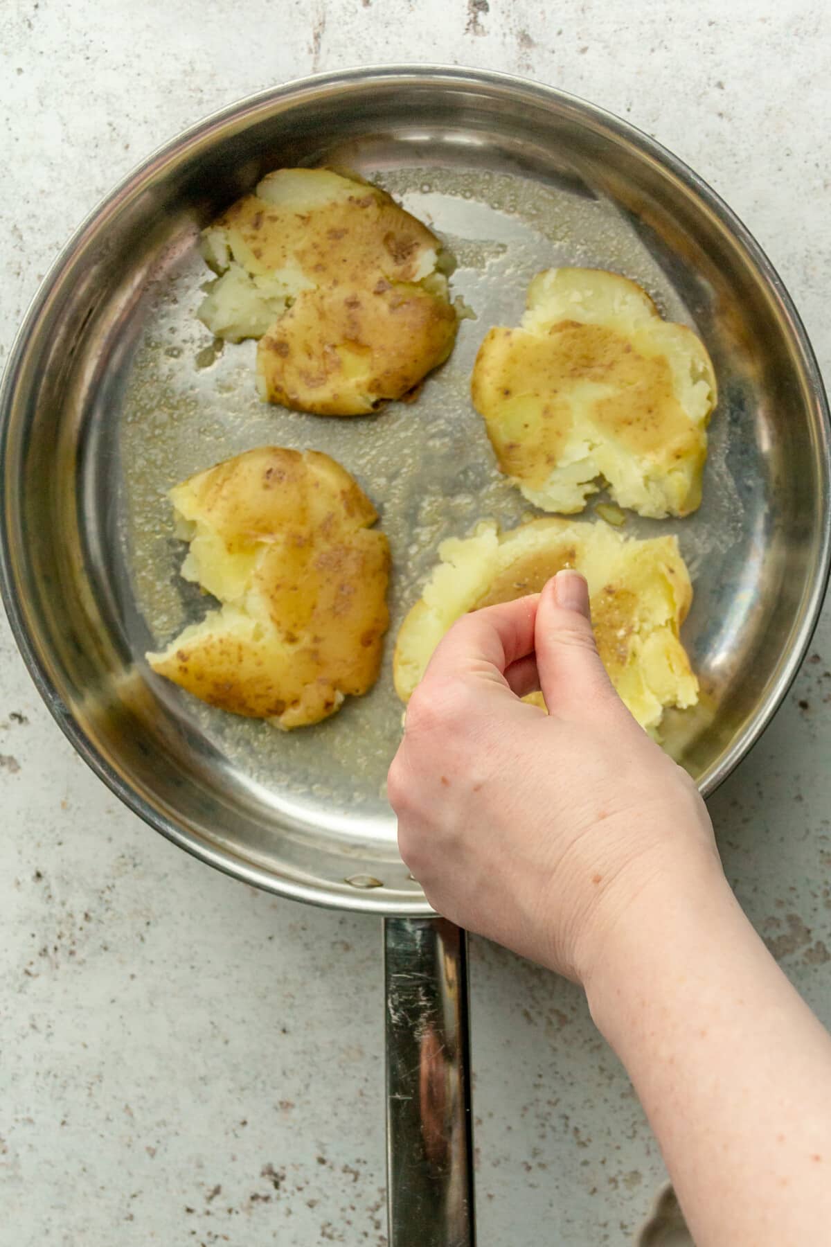 A spice mixture is sprinkled over smashed potatoes in a stainless steel saucepan on a light gray surface.