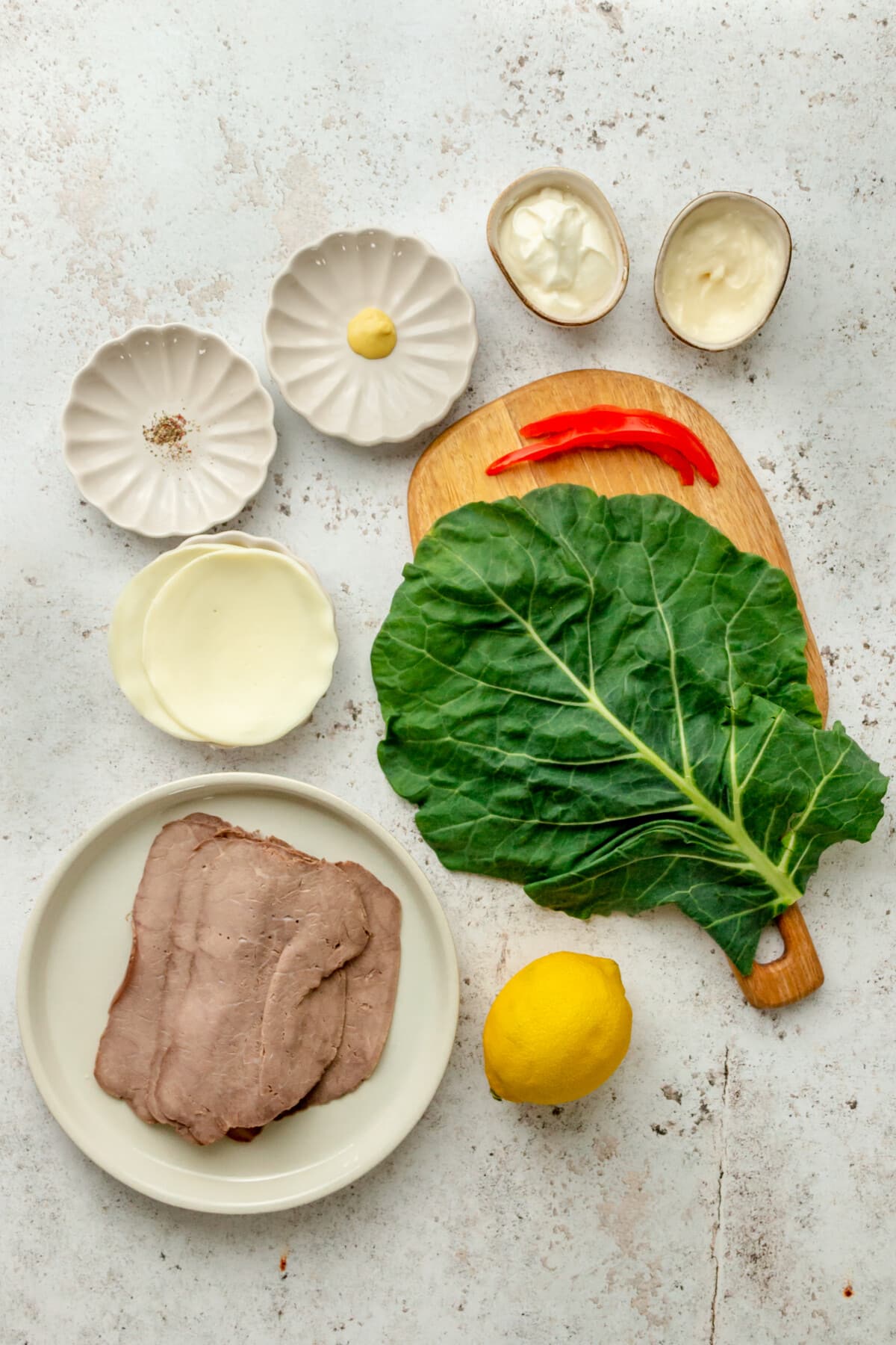 Ingredients for roast beef wraps with horseradish sauce sit in a variety of bowls and plates on a light grey surface.