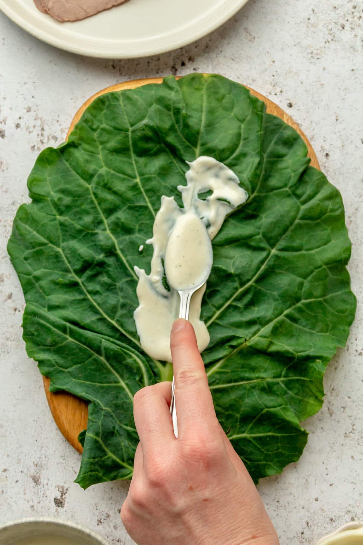 A horseradish sauce is smeared over a collard green leaf on a wood board on a light grey surface.