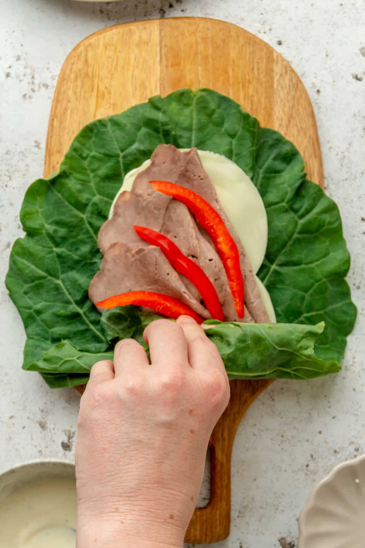 A roast beef wrap is rolled in a collard green leaf, sitting on a wooden board on a light grey surface.