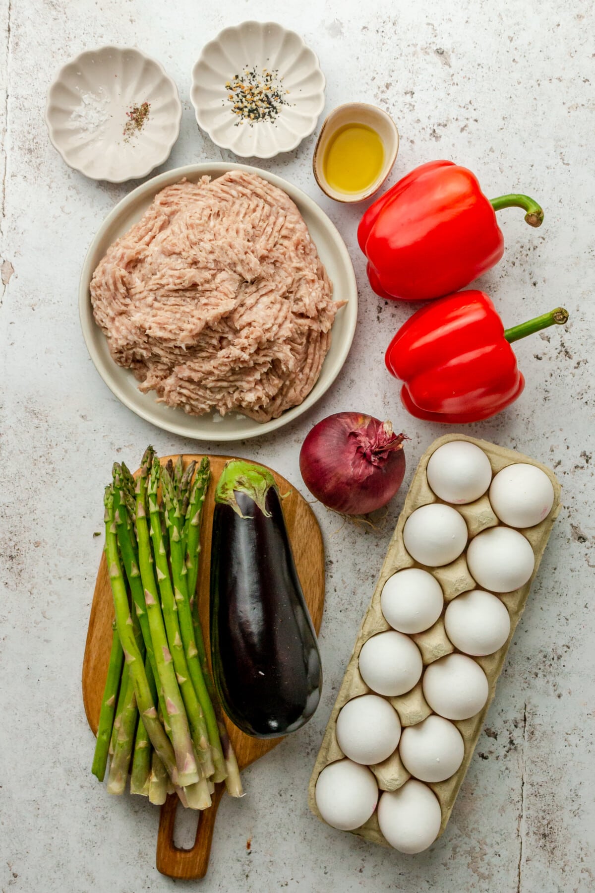 Ingredients for roasted veggie breakfast casserole sit in a variety of bowls and plates on a light grey surface.