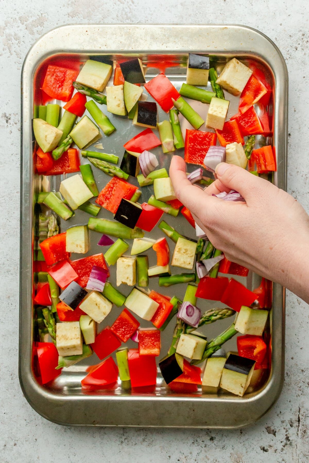 Chopped vegetables are tossed onto a rimmed stainless steel baking sheet on a light grey surface.