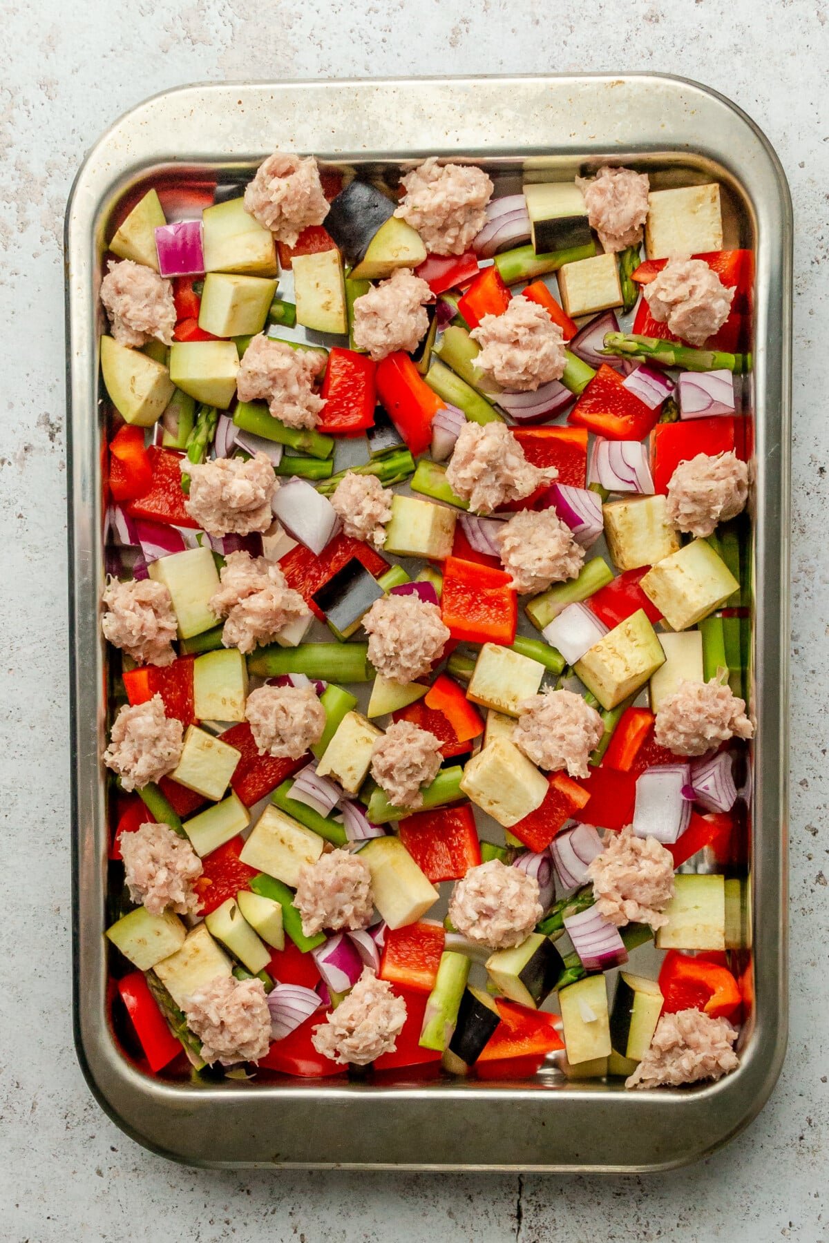 Chopped vegetables and sausage sit on a rimmed stainless steel baking sheet on a light grey surface.