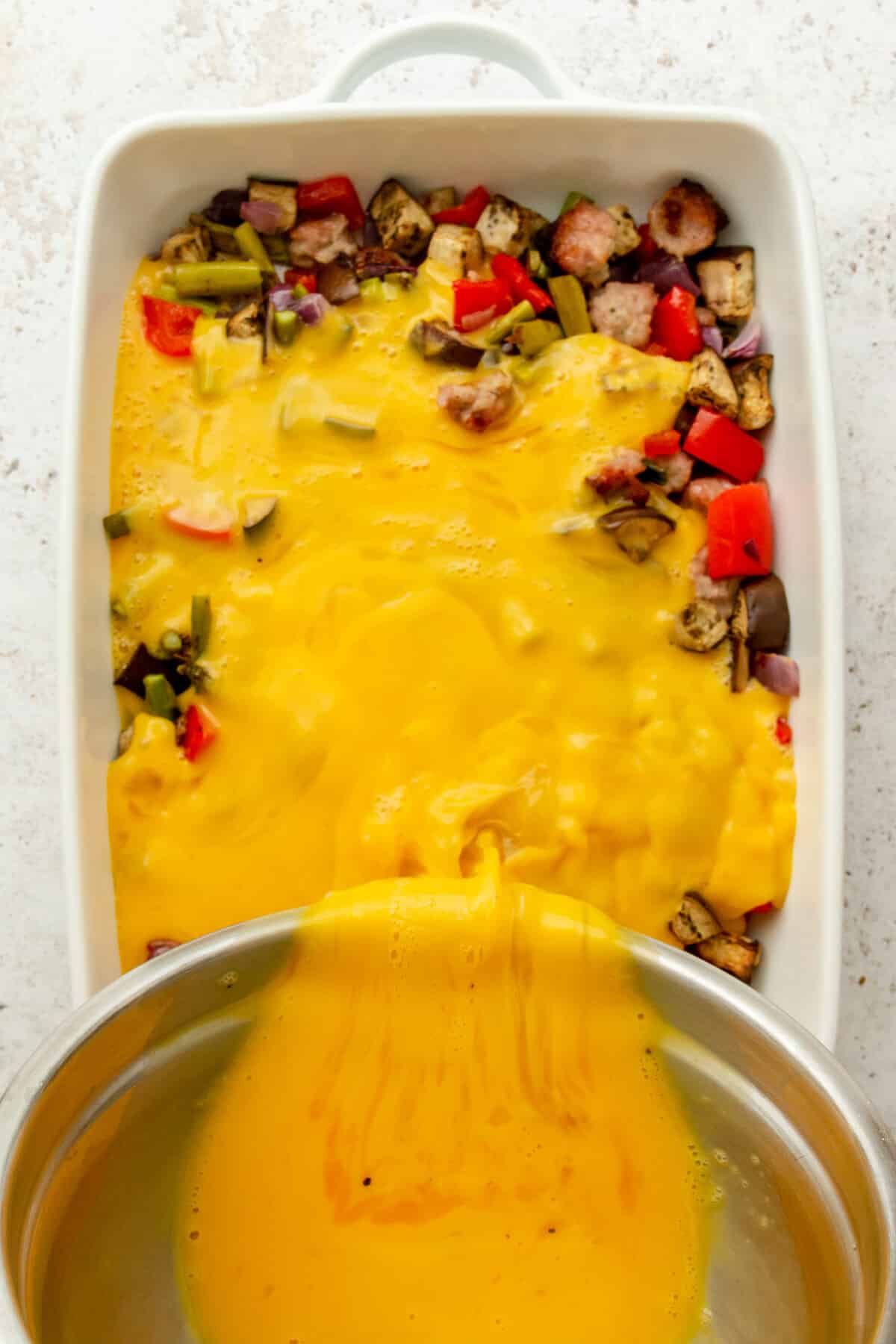 Scrambled eggs are poured over roasted vegetables and sausage in a white ceramic baking dish on a light grey surface.