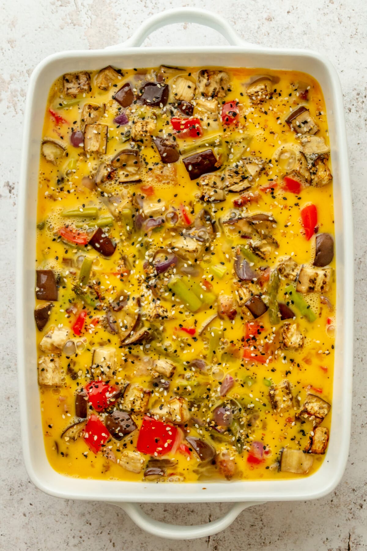 Roasted vegetables, sausage sit in scrambled eggs prior to baking on a light grey surface.