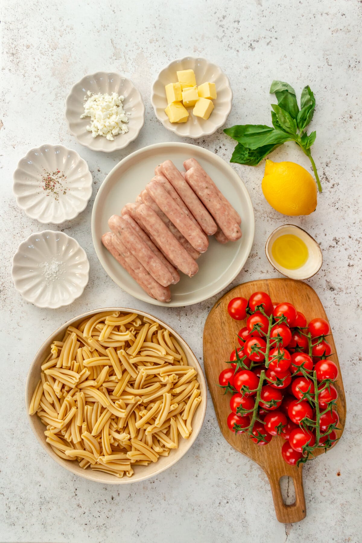 Ingredients for sausage roasted tomato pasta sit in a variety of bowls and plates on a light grey surface.