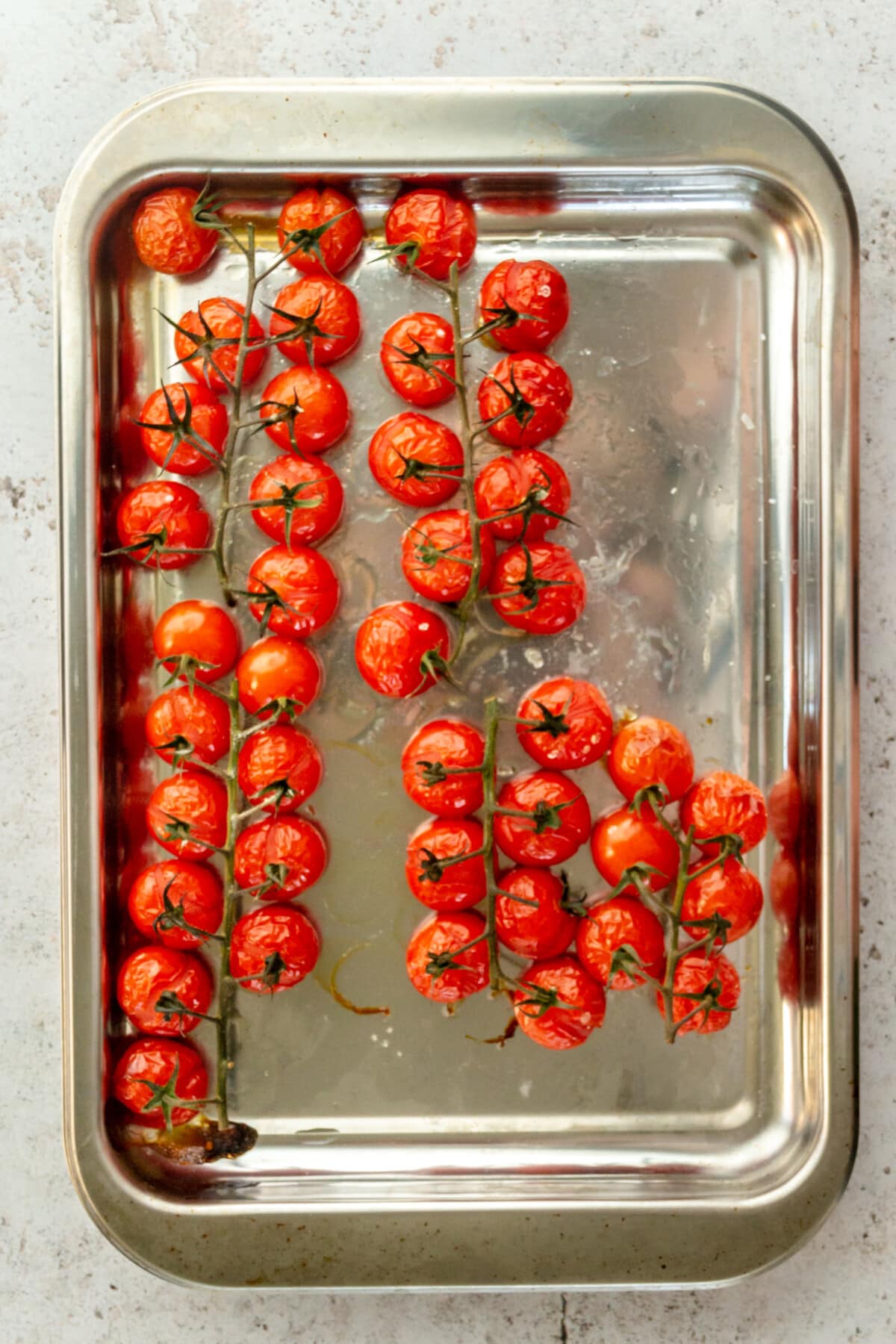 Roasted tomatoes sit on a rimmed stainless steel baking tray on a light grey surface.