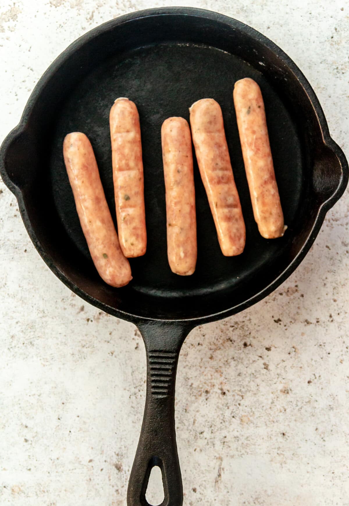 Sausages sit in a cast iron skillet on a light grey surface.