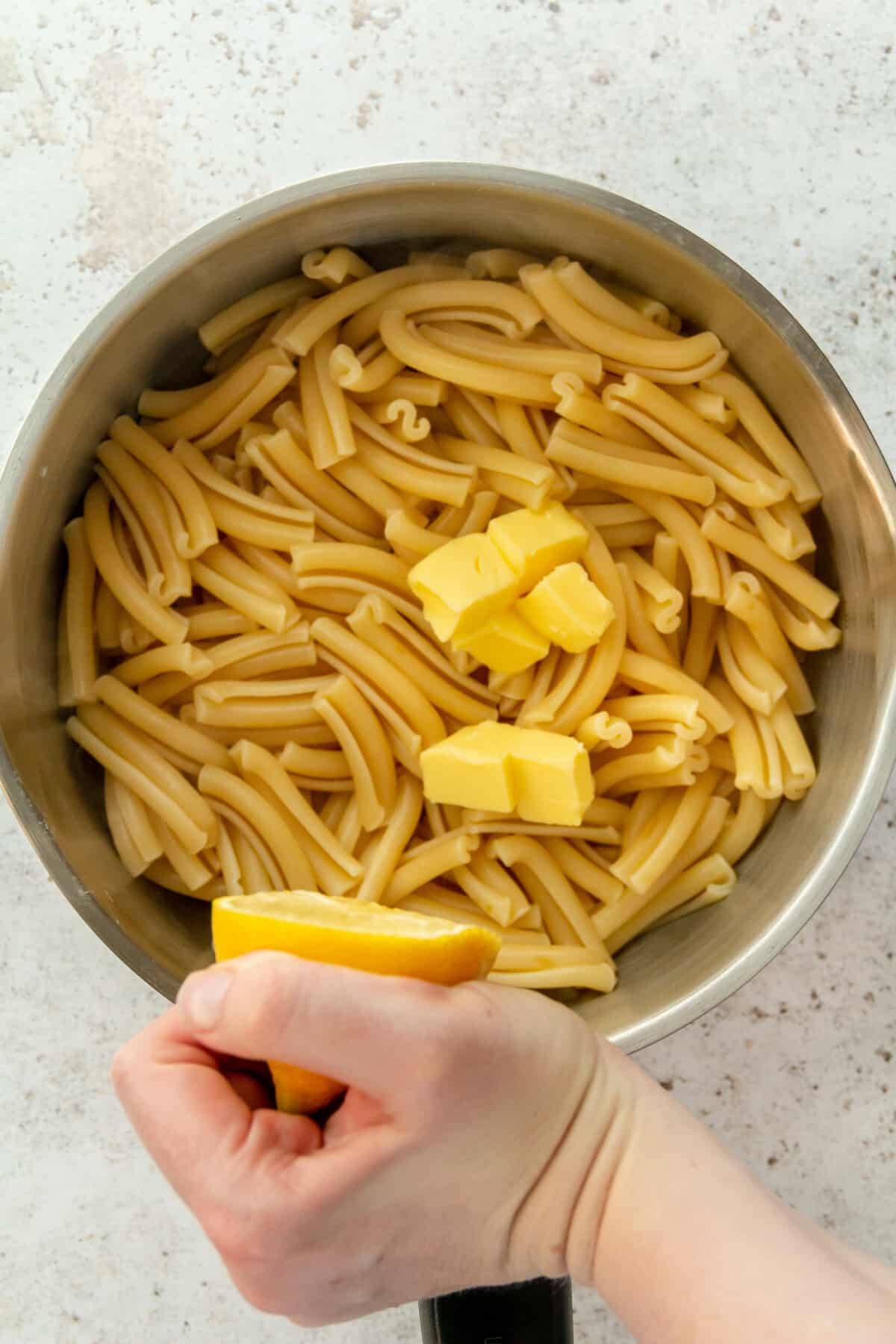 Lemon juice is squeezed over cooked pasta topped with cubes of butter in a stainless steel saucepan on a light grey surface.