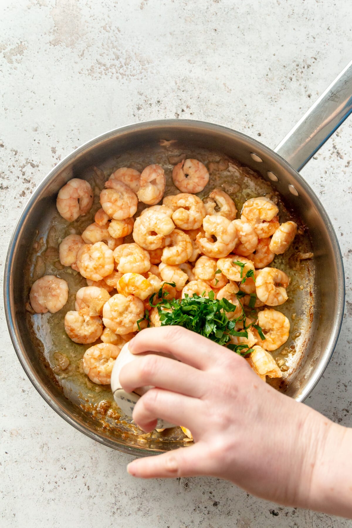 A hand is shown pouring a bowl of chopped cilantro over the top of the shrimp in the pan.