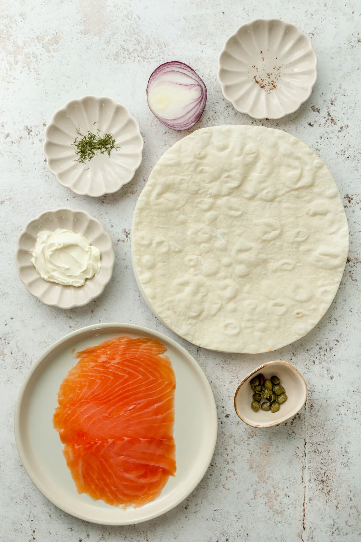 Ingredients for smoked salmon lunch wraps sit in a variety of bowls and plates on a light grey surface.