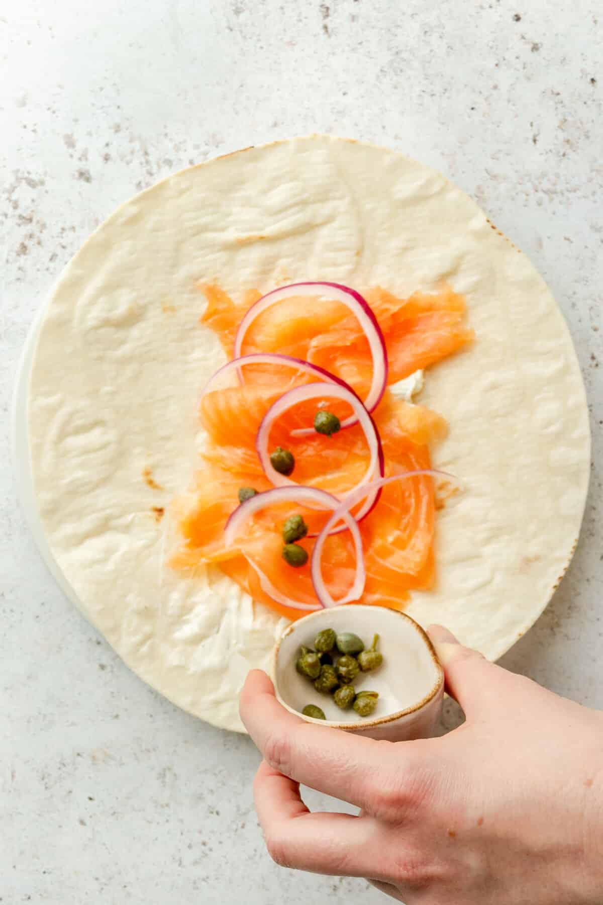 Capers are tossed over pieces of smoked salmon and slices of red onion on a wrap on a light grey surface.