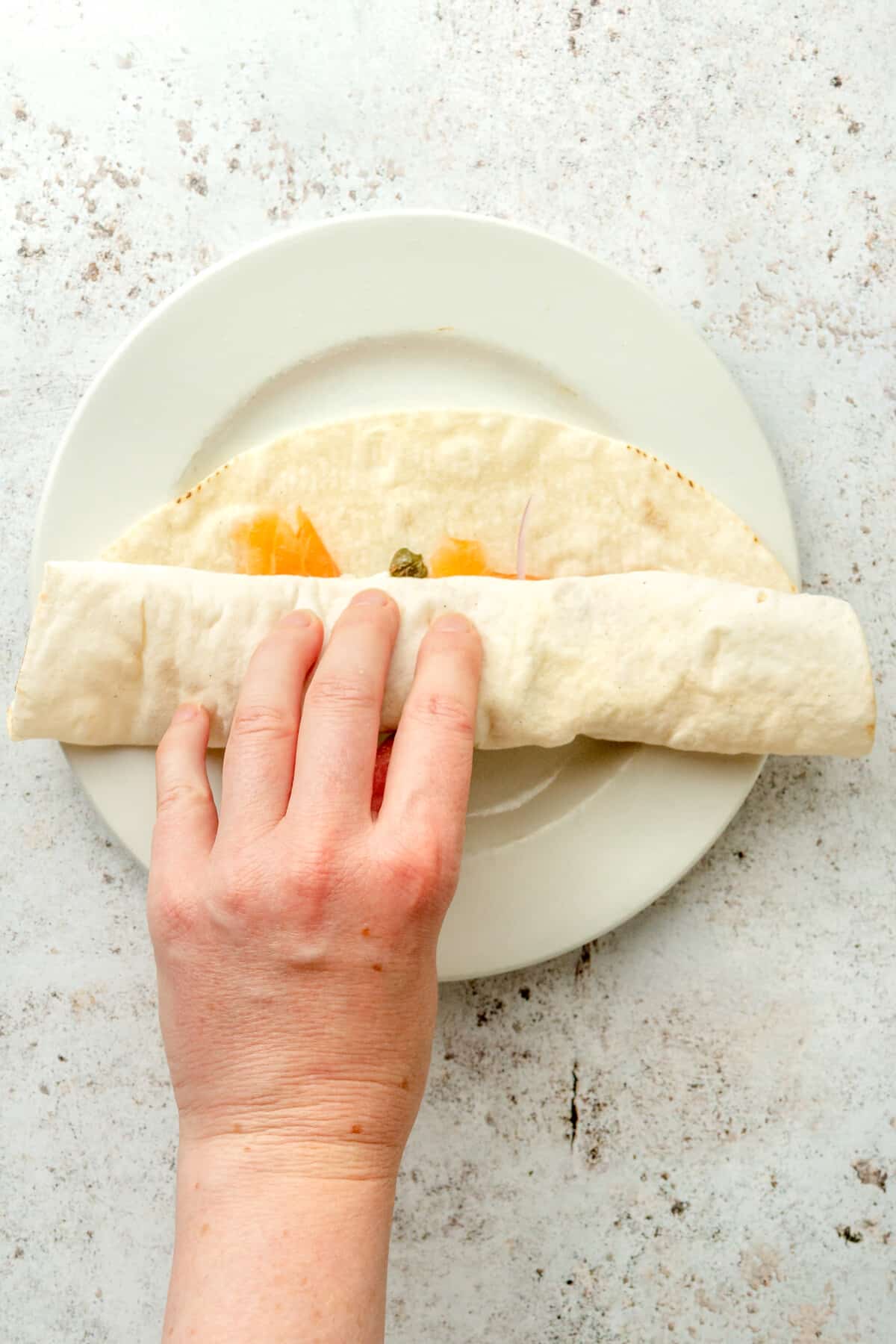 A smoked salmon lunch wrap is rolled on a white plate on a light grey surface.
