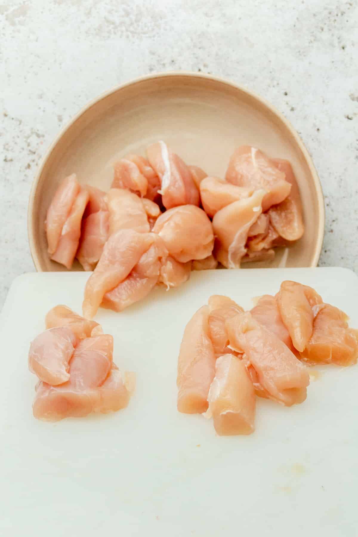 Sliced pieces of chicken tenders are slid into a shallow ceramic bowl on a light grey surface.