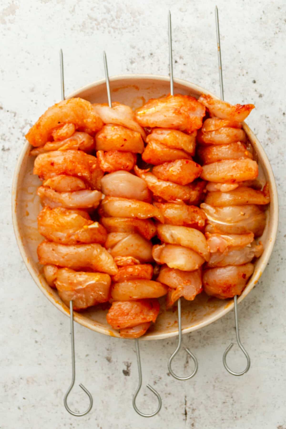 Skewers of spiced chicken tenders sit on top of a shallow ceramic bowl on a light grey surface.