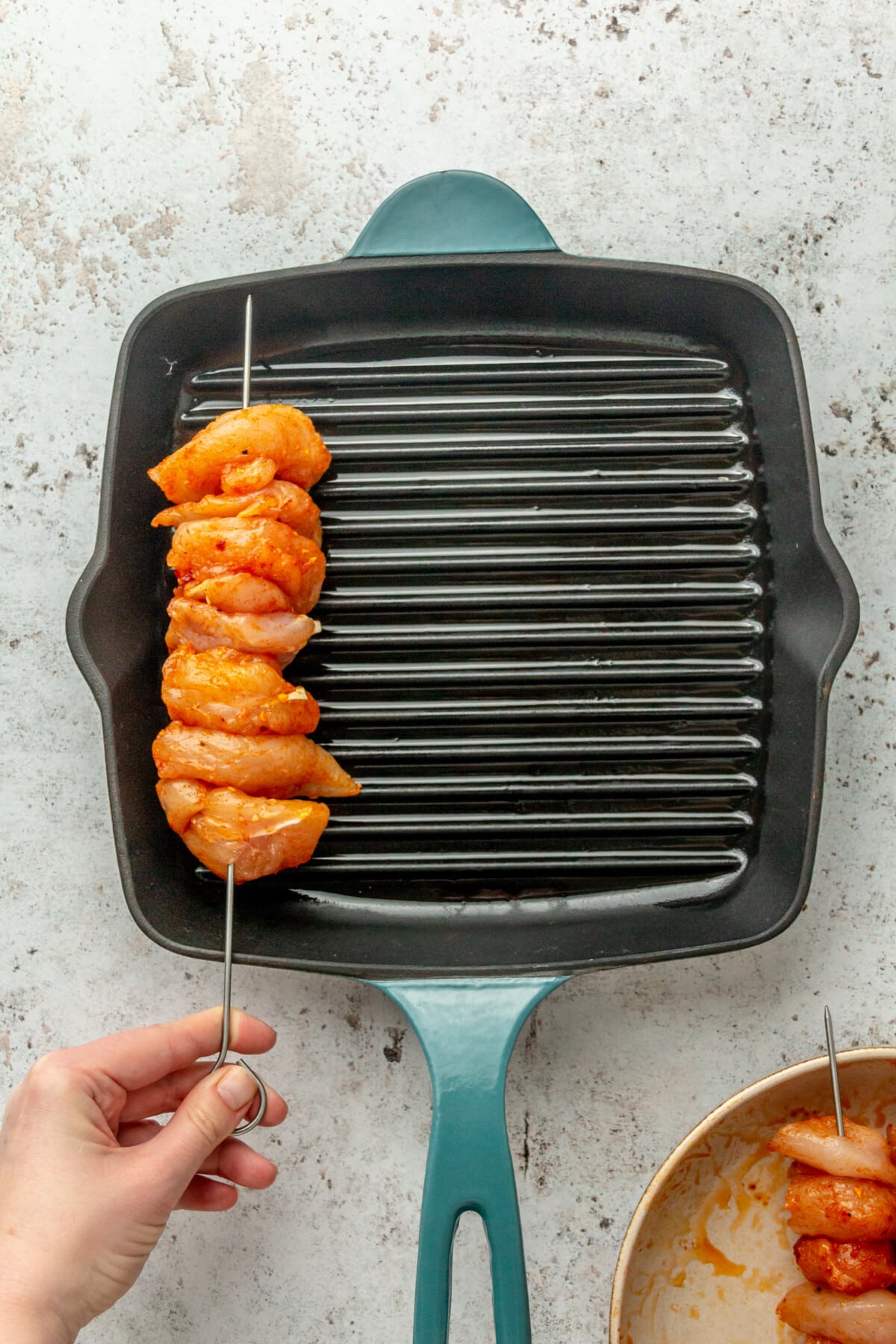 A skewer of spiced chicken tenders is placed into a grill cast iron skillet on a light grey surface.